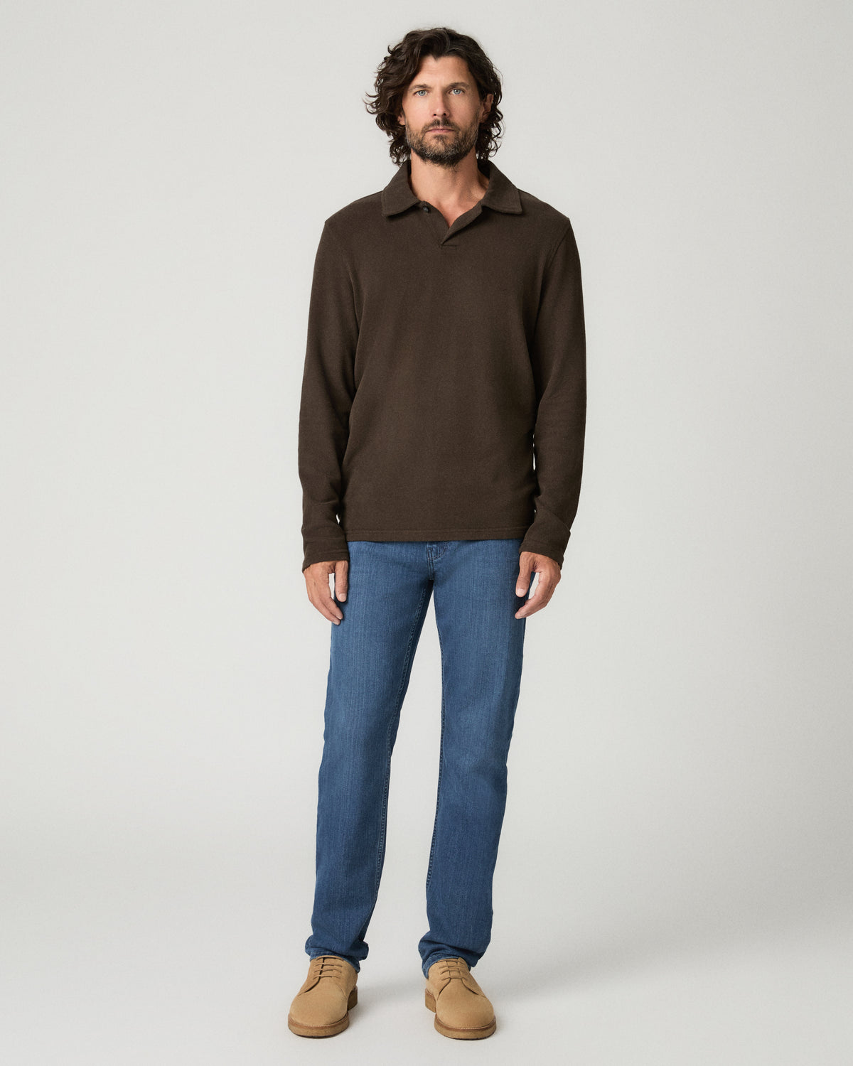 Full-length front view of a man with dark curly hair wearing a dark brown long-sleeve popover polo shirt, paired with medium blue slim-straight jeans and tan suede shoes.