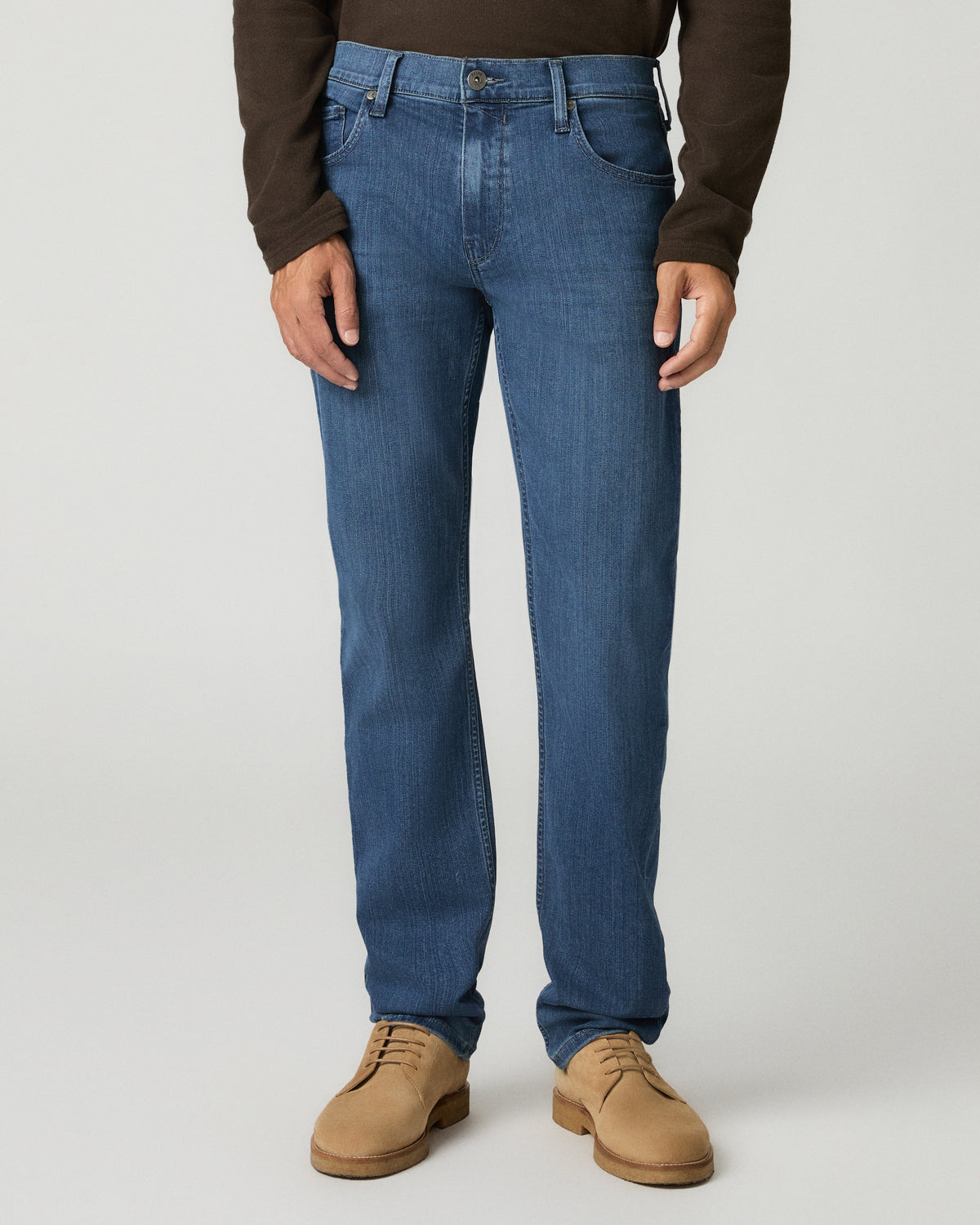Close-up front view of a man wearing medium blue slim-straight jeans, dark brown sleeve visible above and tan suede shoes below.