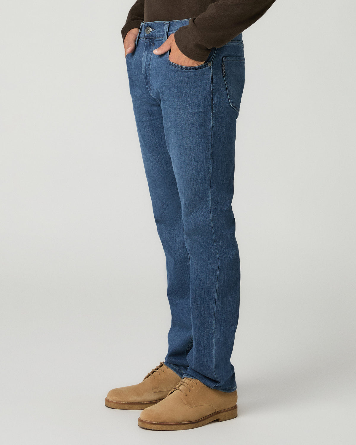 Side profile view of a man with hands in his pockets wearing medium blue slim-straight jeans and tan suede shoes.