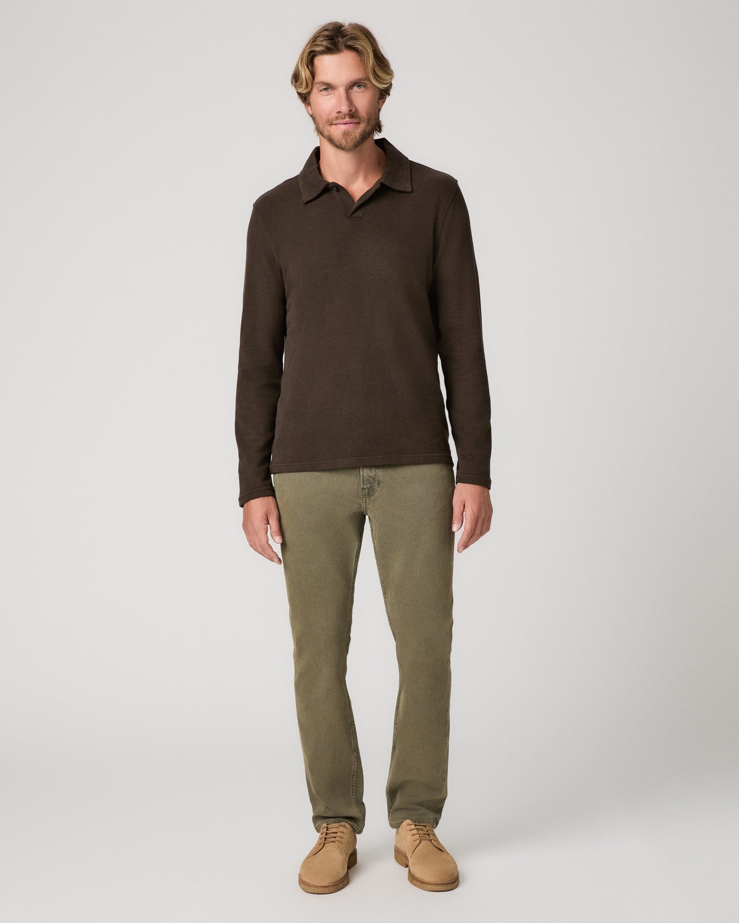 A man wearing a brown long sleeve shirt and olive green pant paired with brown boots standing against a light gray studio background.