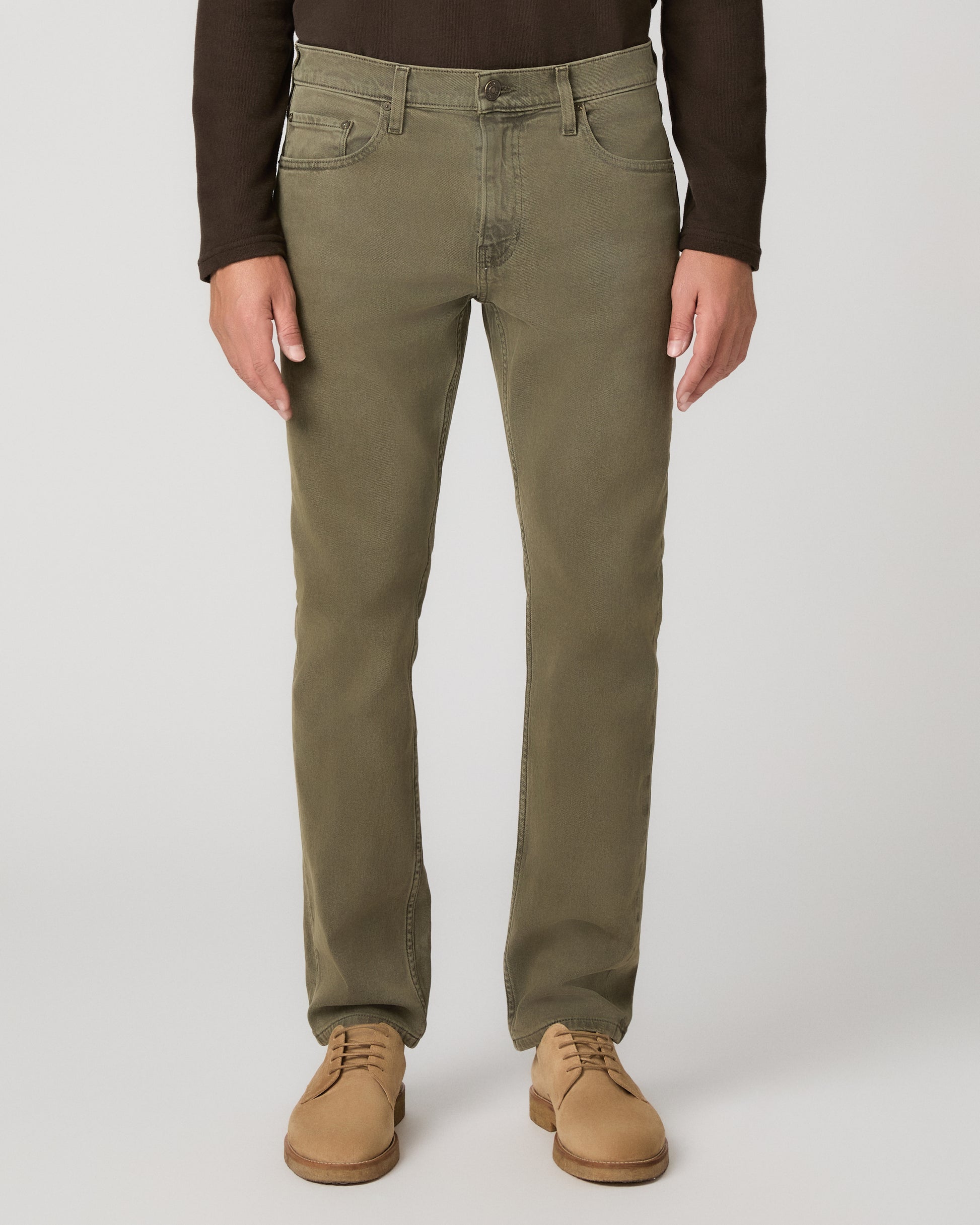 A man wearing a brown long sleeve shirt and olive green pant paired with brown boots standing against a light gray studio background.