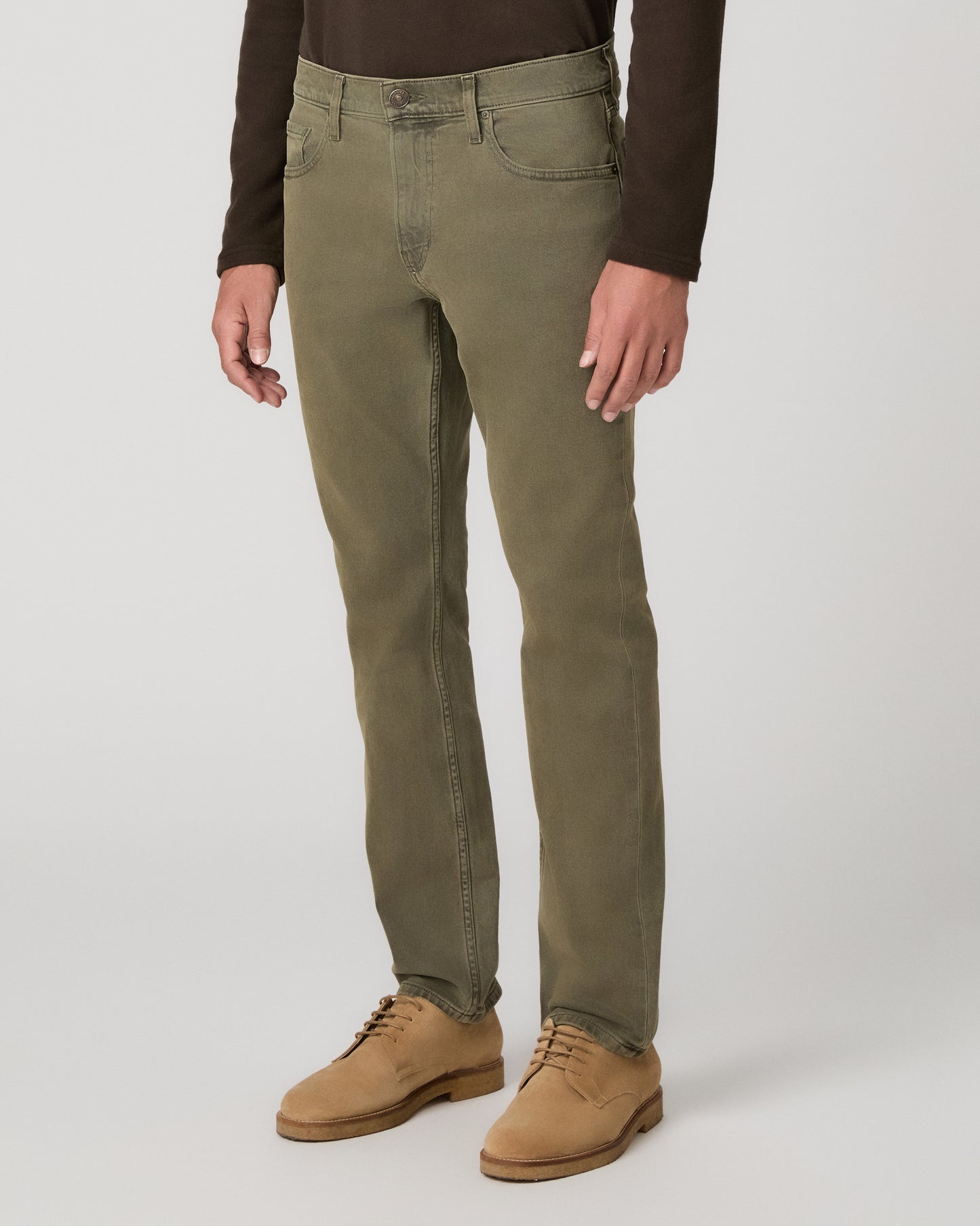A man wearing a brown long sleeve shirt and olive green pant paired with brown boots standing against a light gray studio background.