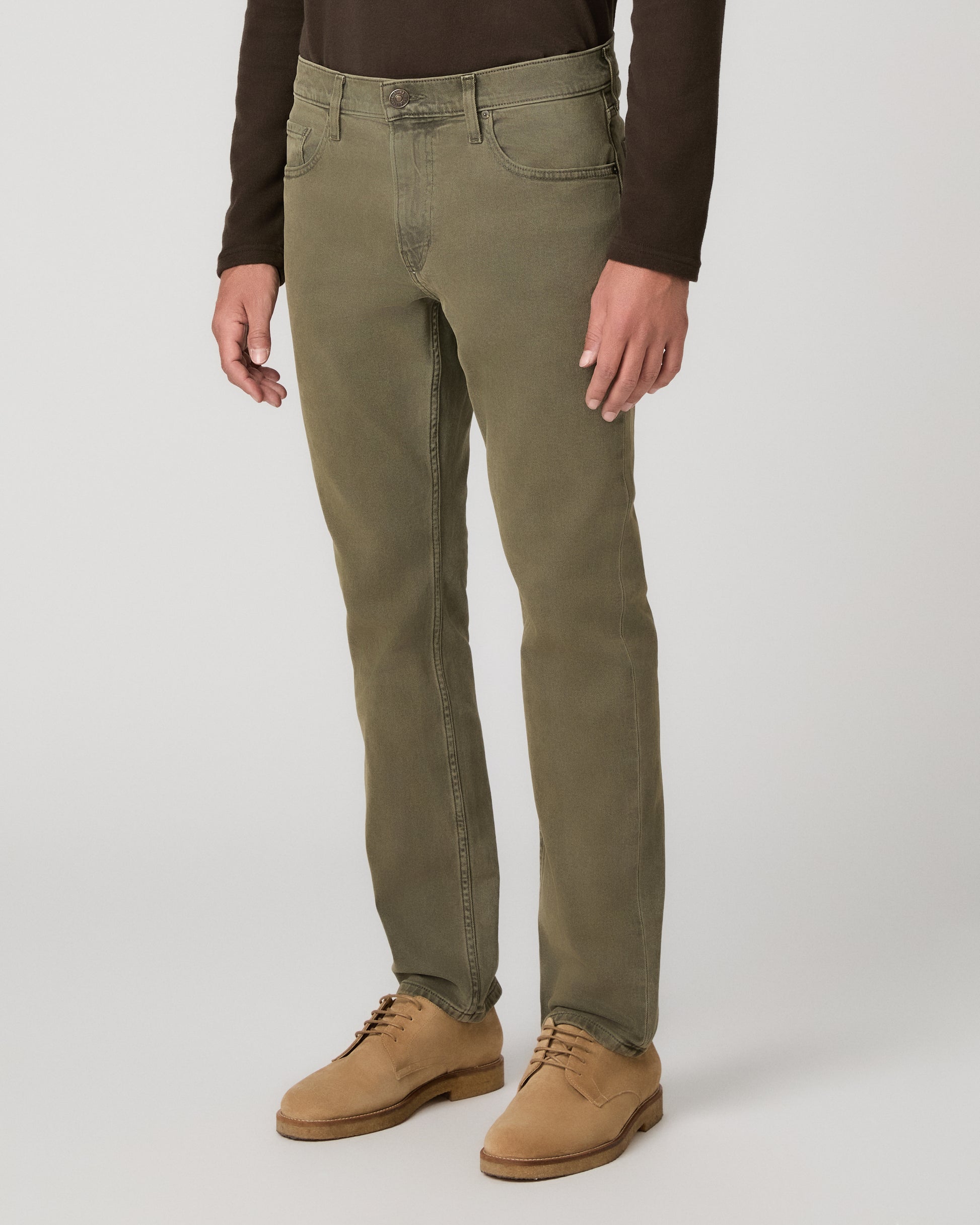A man wearing a brown long sleeve shirt and olive green pant paired with brown boots standing against a light gray studio background.