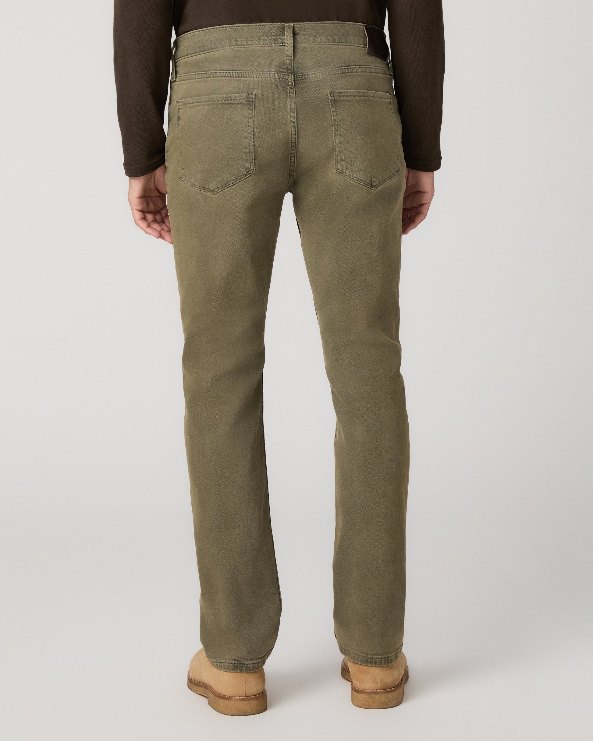 A man wearing a brown long sleeve shirt and olive green pant paired with brown boots standing against a light gray studio background.