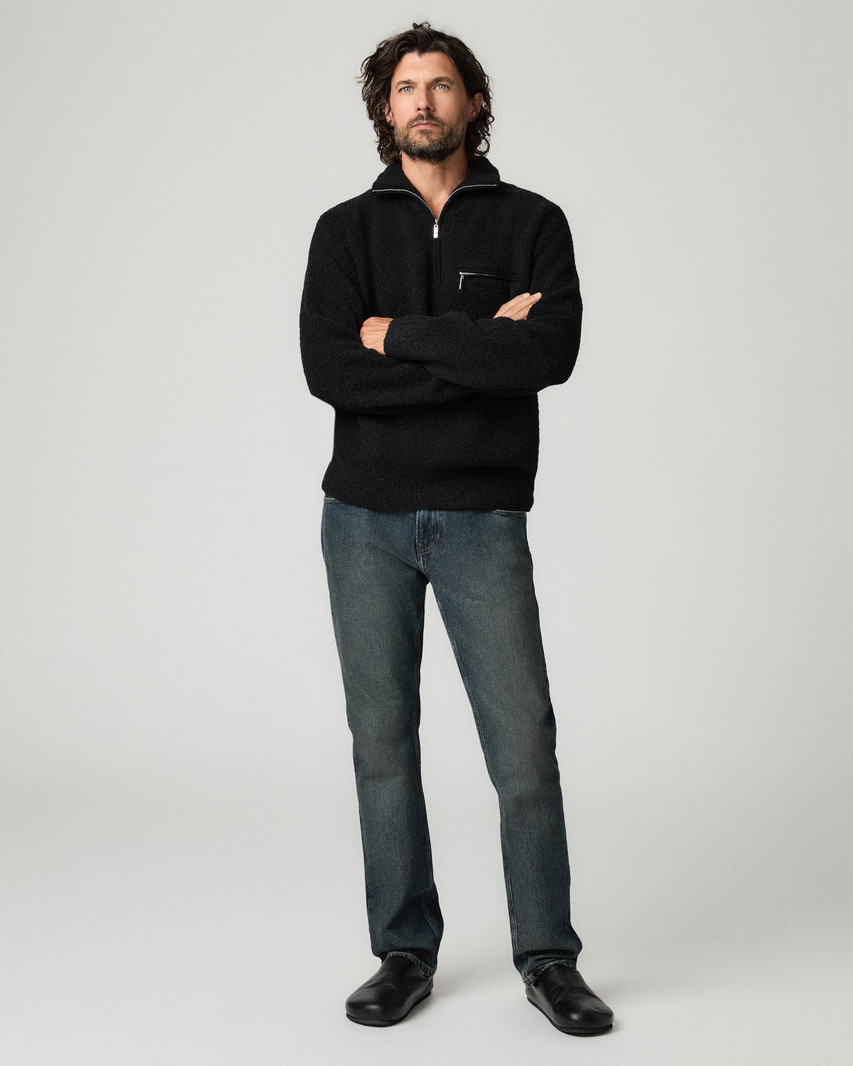 Full-length front view of a man with dark curly hair wearing a black quarter-zip sweater with arms crossed, paired with charcoal slim-straight jeans and black leather shoes.