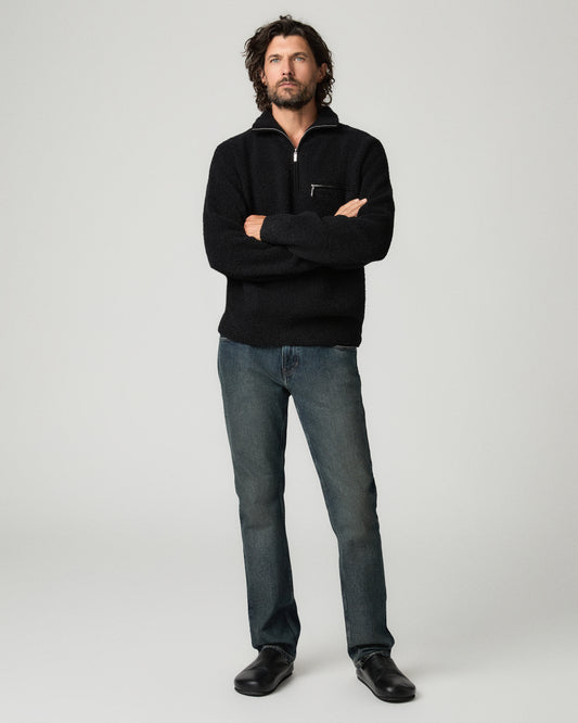 Full-length front view of a man with dark curly hair wearing a black quarter-zip sweater with arms crossed, paired with charcoal slim-straight jeans and black leather shoes.