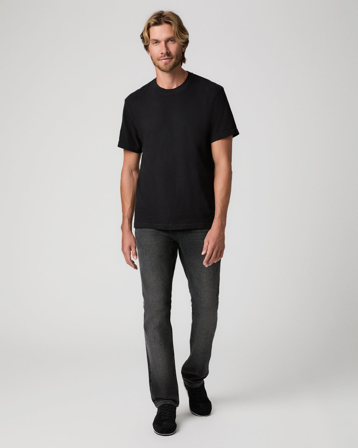 A man wearing a plain black short-sleeve crewneck t-shirt, dark gray slim-fit jeans, and black sneakers, standing against a light gray studio background.