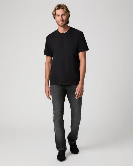 A man wearing a plain black short-sleeve crewneck t-shirt, dark gray slim-fit jeans, and black sneakers, standing against a light gray studio background.