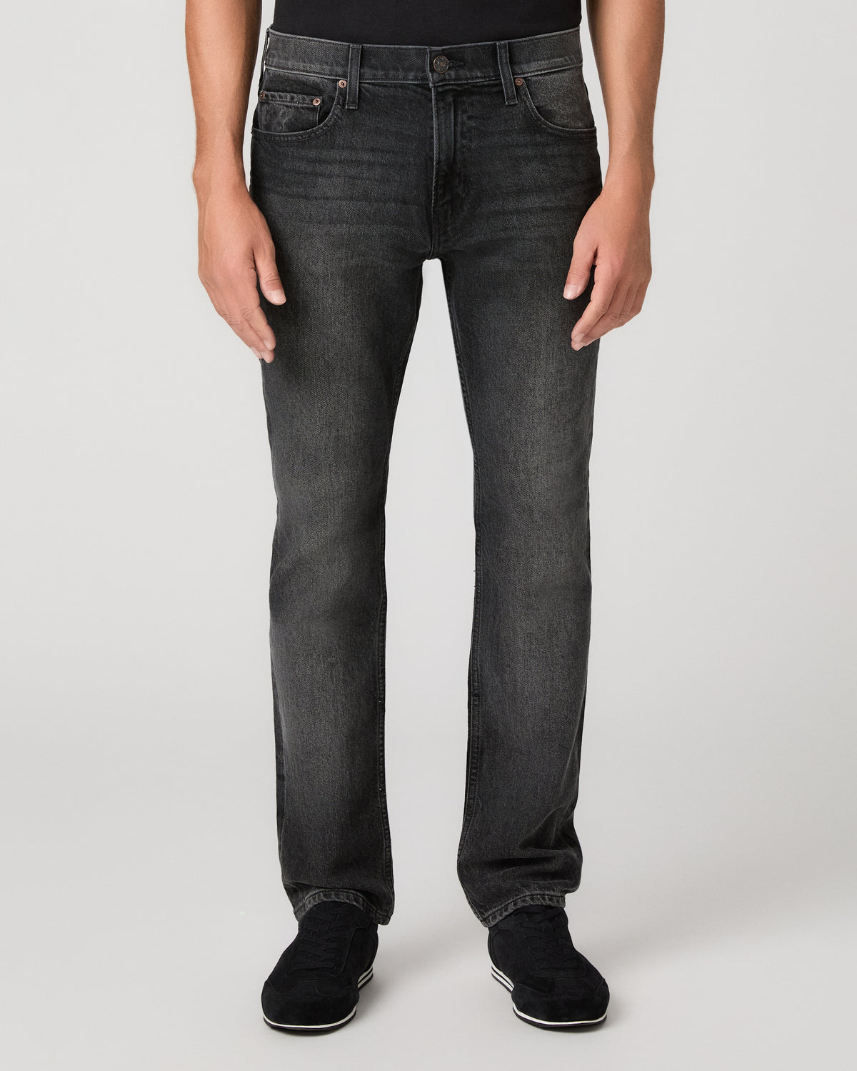 A man wearing a plain black short-sleeve crewneck t-shirt, dark gray slim-fit jeans, and black sneakers, standing against a light gray studio background.