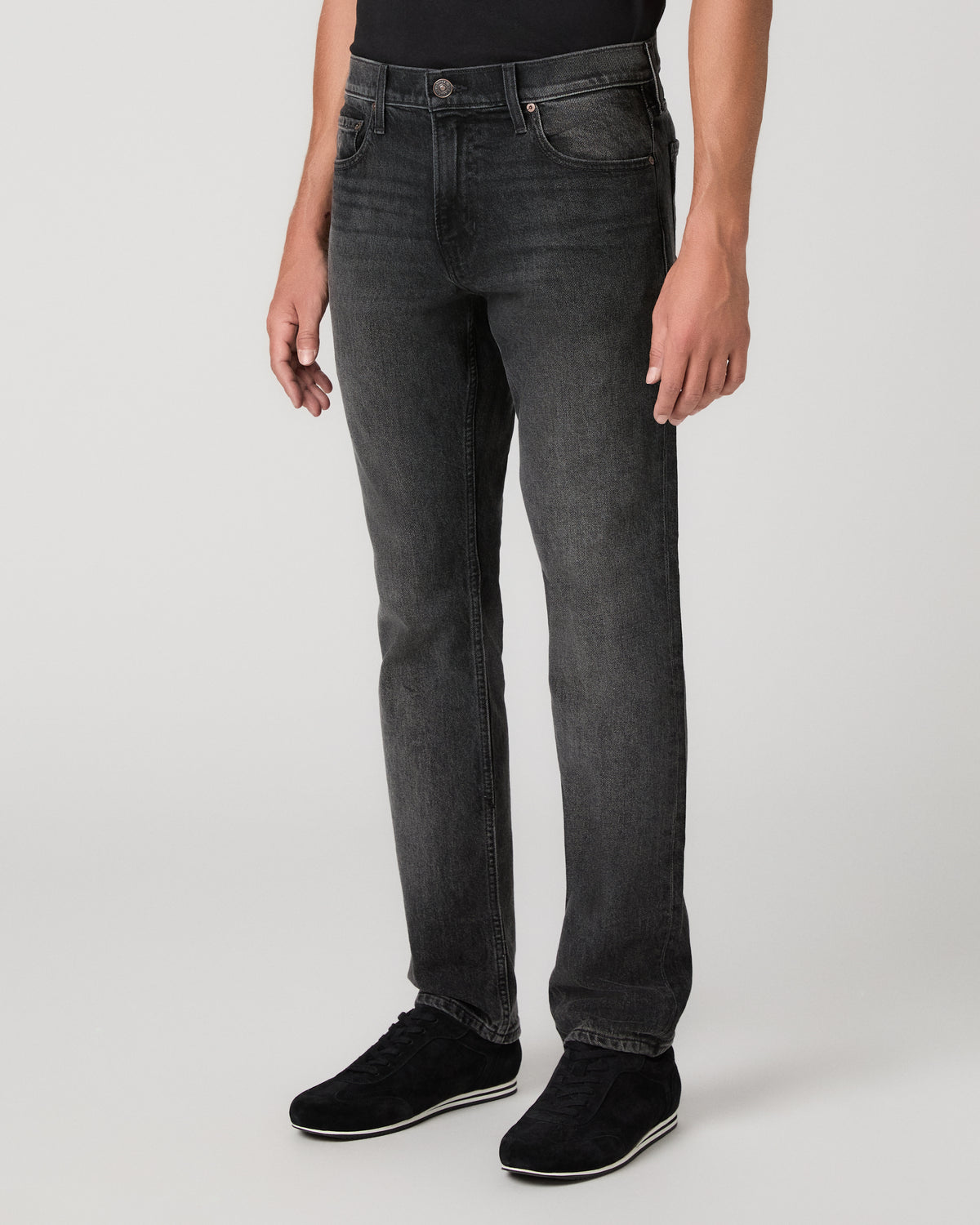 A man wearing a plain black short-sleeve crewneck t-shirt, dark gray slim-fit jeans, and black sneakers, standing against a light gray studio background.