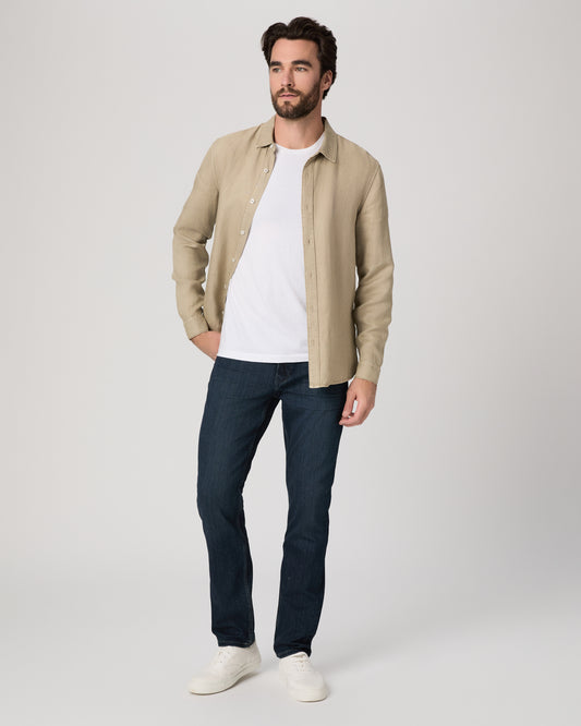 Man wearing a beige shirt, white t-shirt, dark jeans, and white shoes on a white background