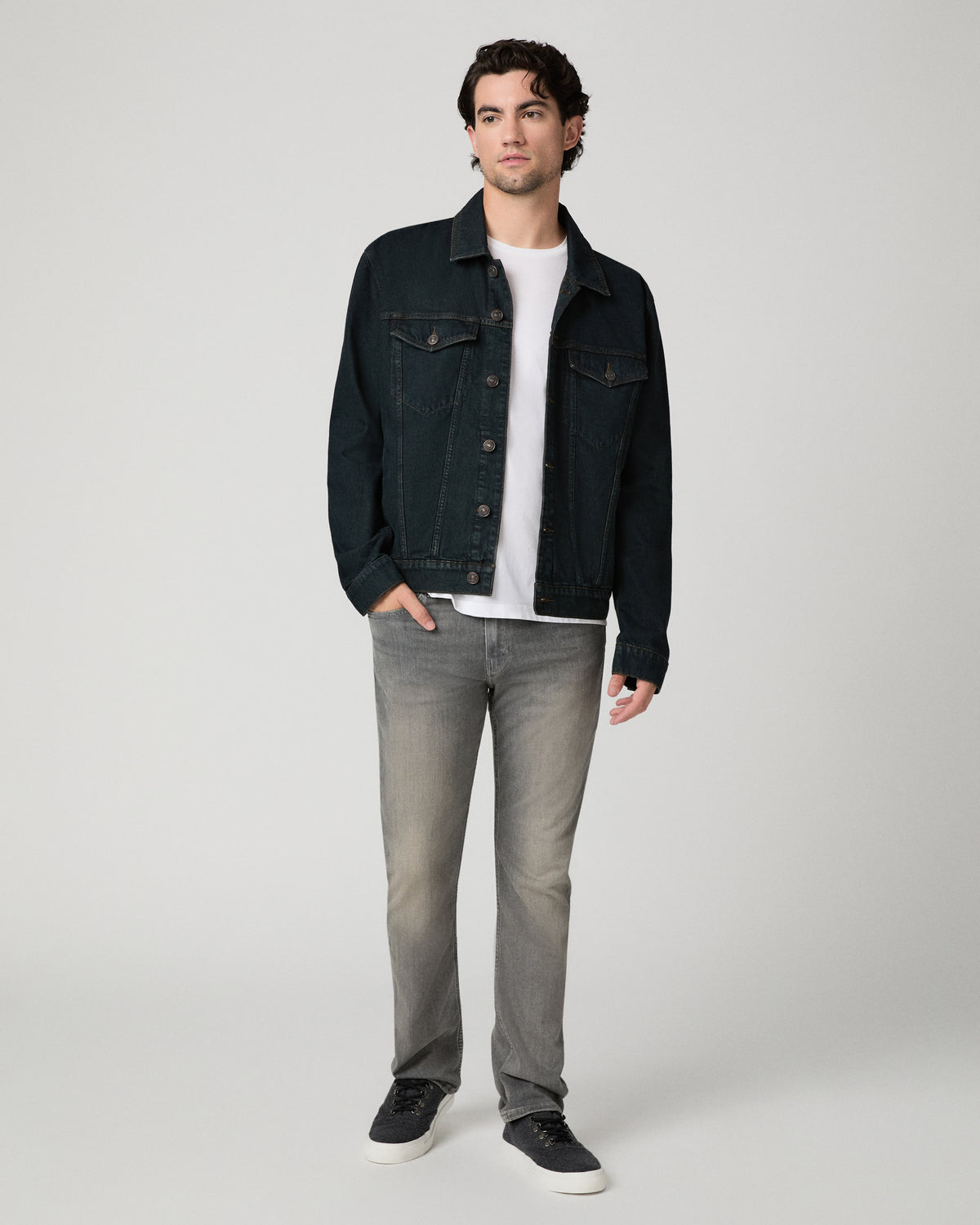 Man wearing white shirt, dark denim jacket, and grey pants against a white background