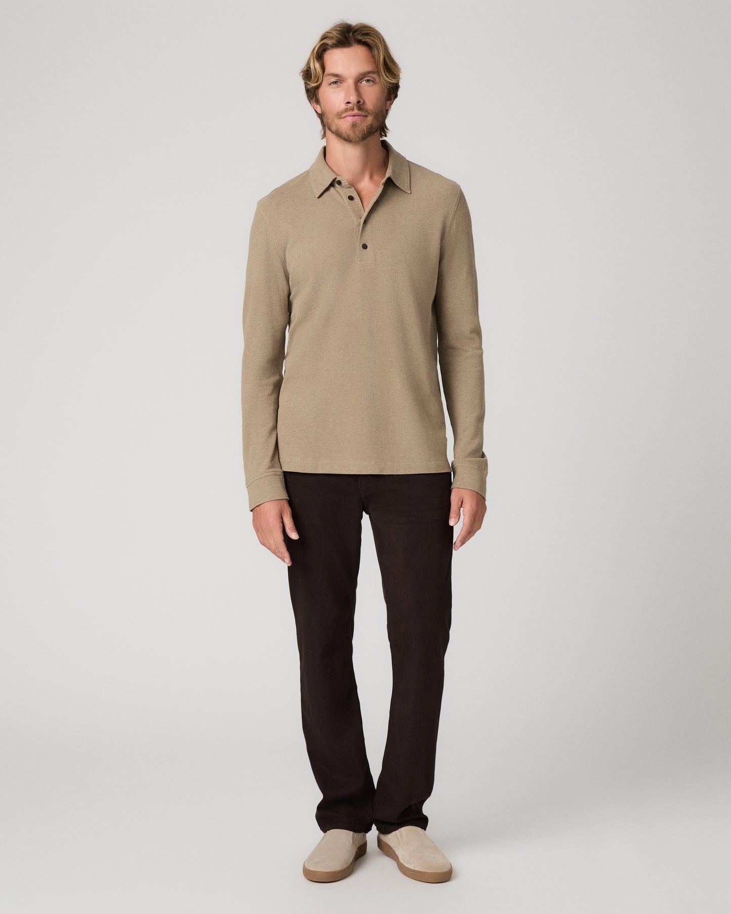 A man modeling a beige long-sleeve polo shirt with dark brown trousers and beige slip-on shoes, standing against a light gray studio background.