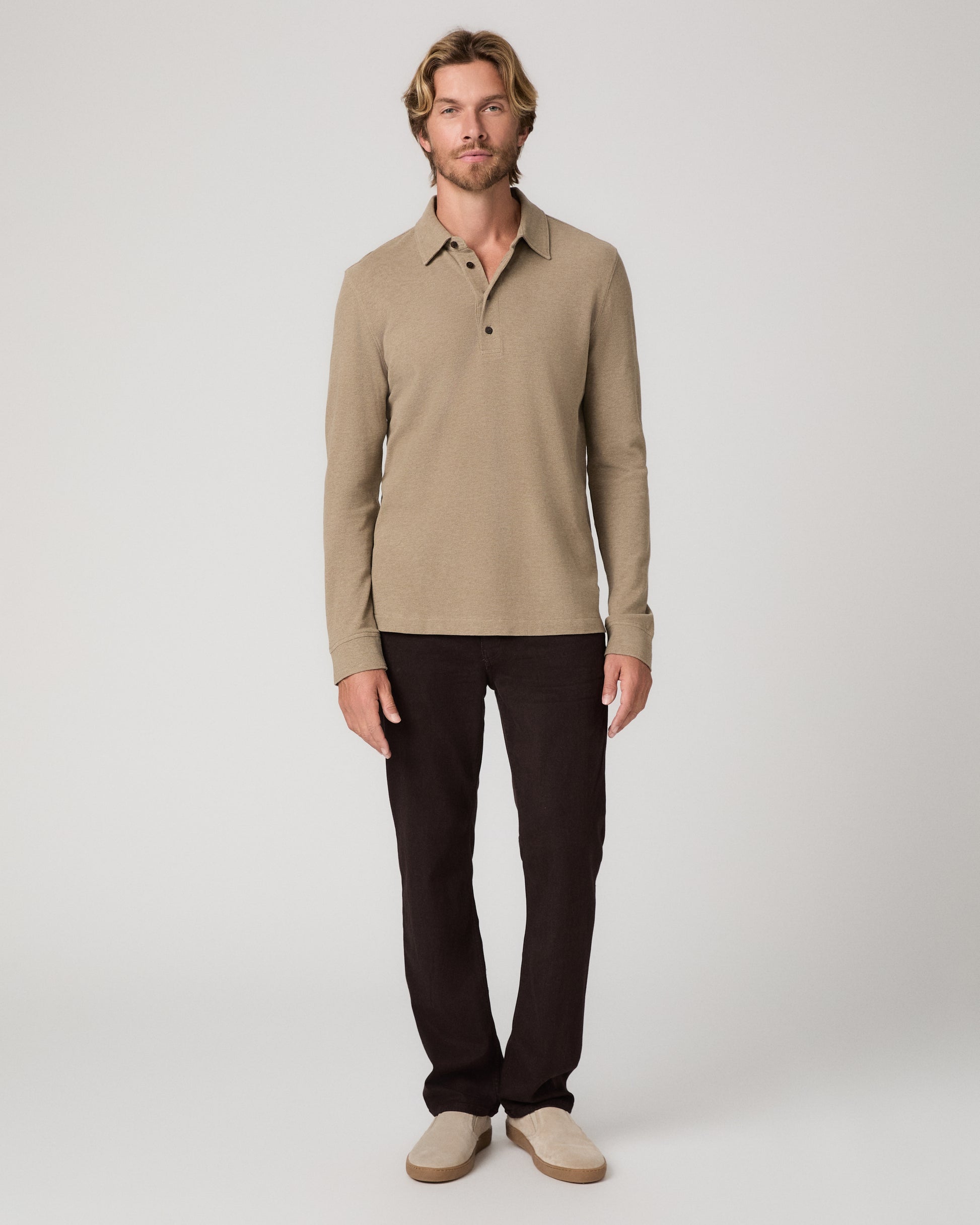 A man modeling a beige long-sleeve polo shirt with dark brown trousers and beige slip-on shoes, standing against a light gray studio background.