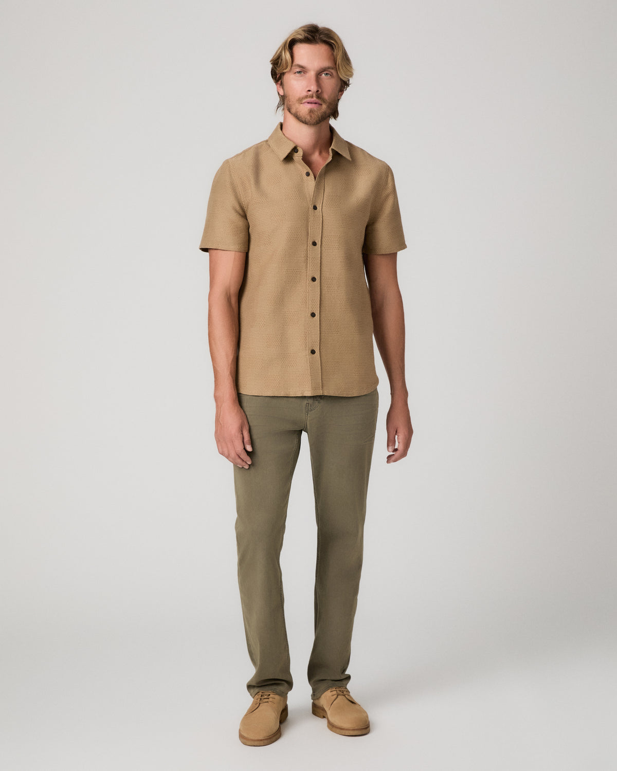 A man modeling a short-sleeve button-up shirt in tan and olive green pants, standing against a light gray studio background.