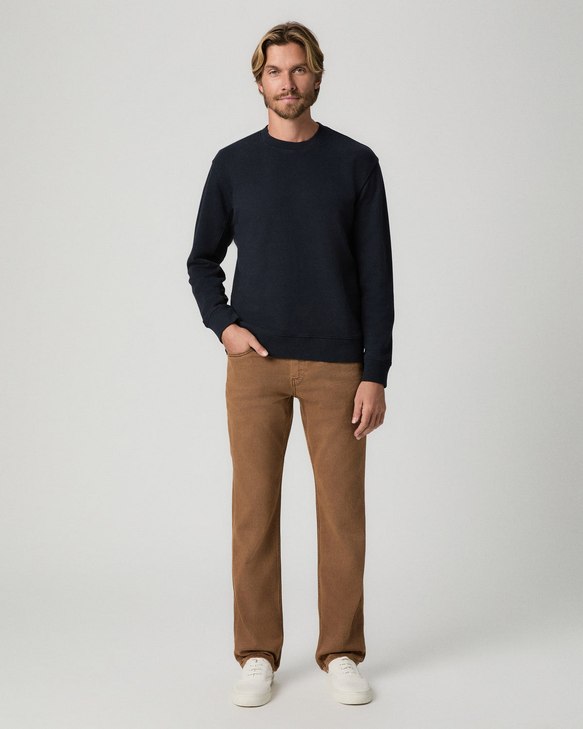Full-length view of model with one hand in his pocket, wearing a navy crewneck sweater with the Normandie 32 Inch Straight Leg Jean in Vintage Sierra Canyon and white sneakers