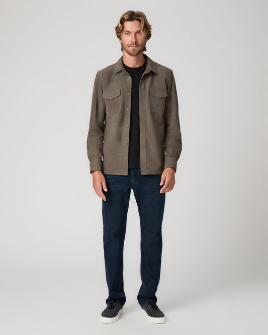 A man modeling a brown suede button-up shirt jacket over a black t-shirt, dark blue jeans, and black sneakers, photographed against a neutral light gray studio background.
