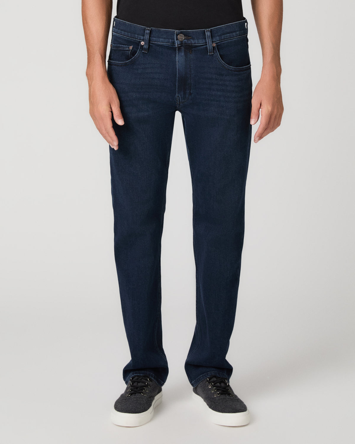 A man modeling a black t-shirt, dark blue jeans, and black sneakers, photographed against a neutral light gray studio background.
