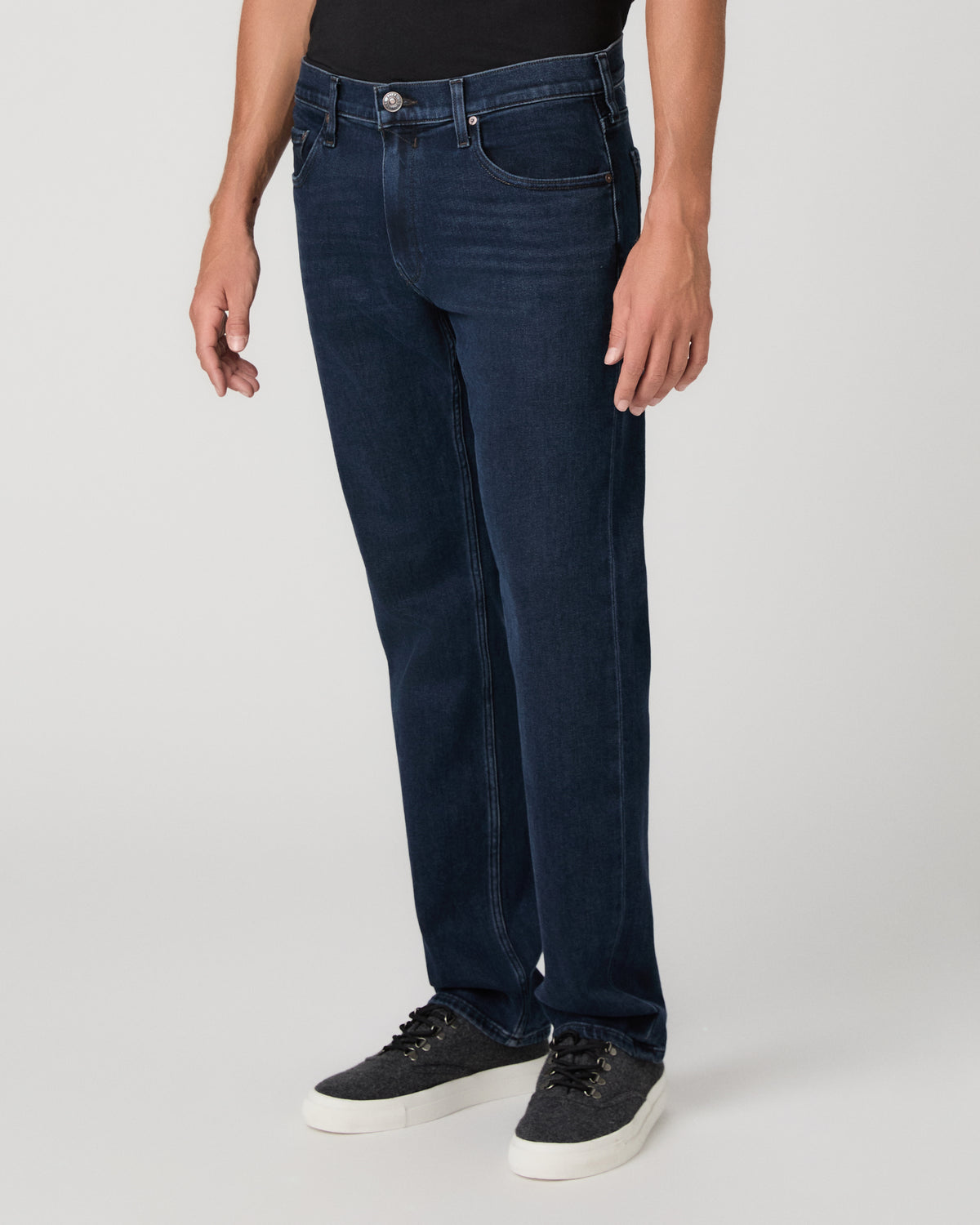 A man modeling a black t-shirt, dark blue jeans, and black sneakers, photographed against a neutral light gray studio background.