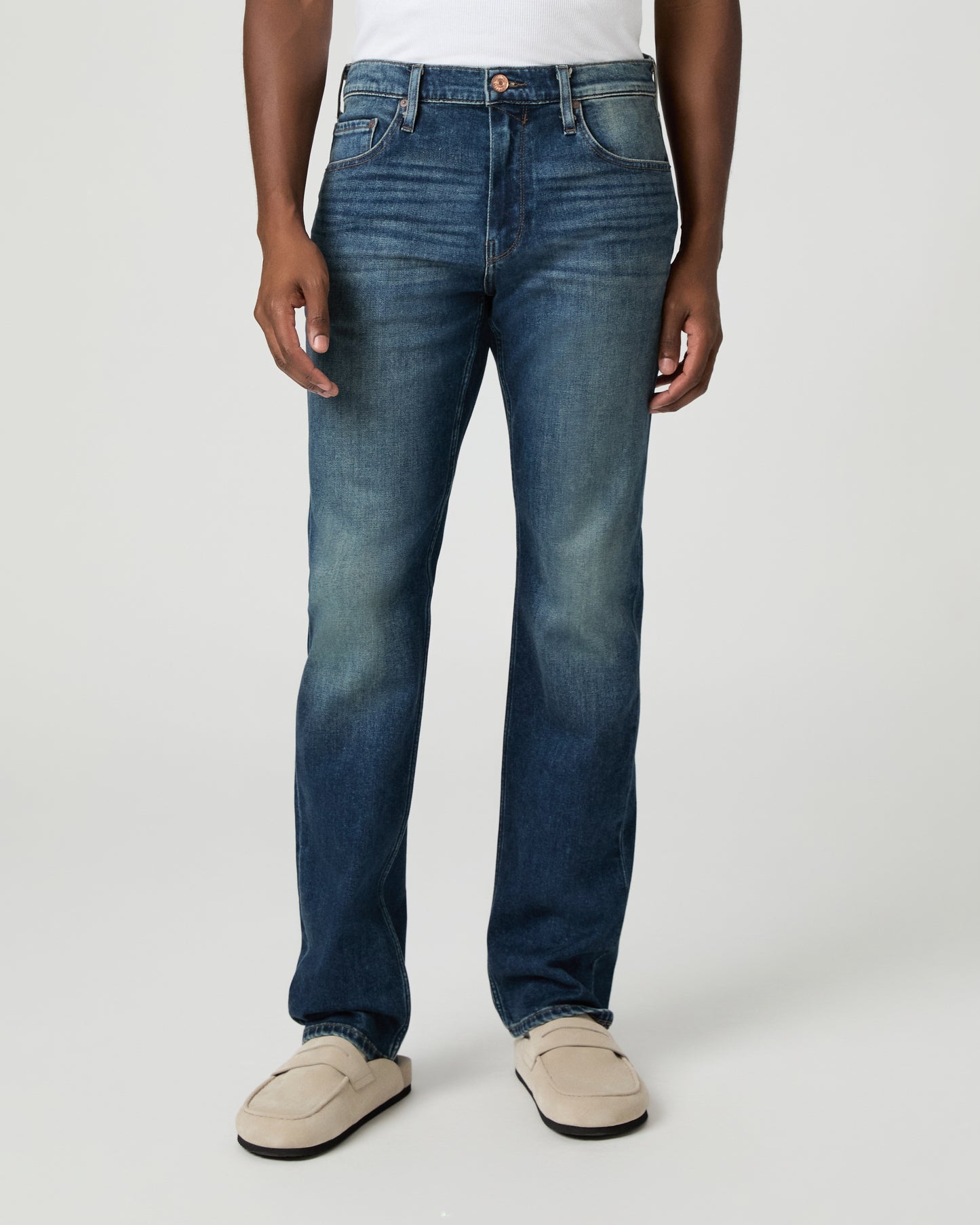 Man wearing a white shirt, blue jeans, and beige shoes on a plain background