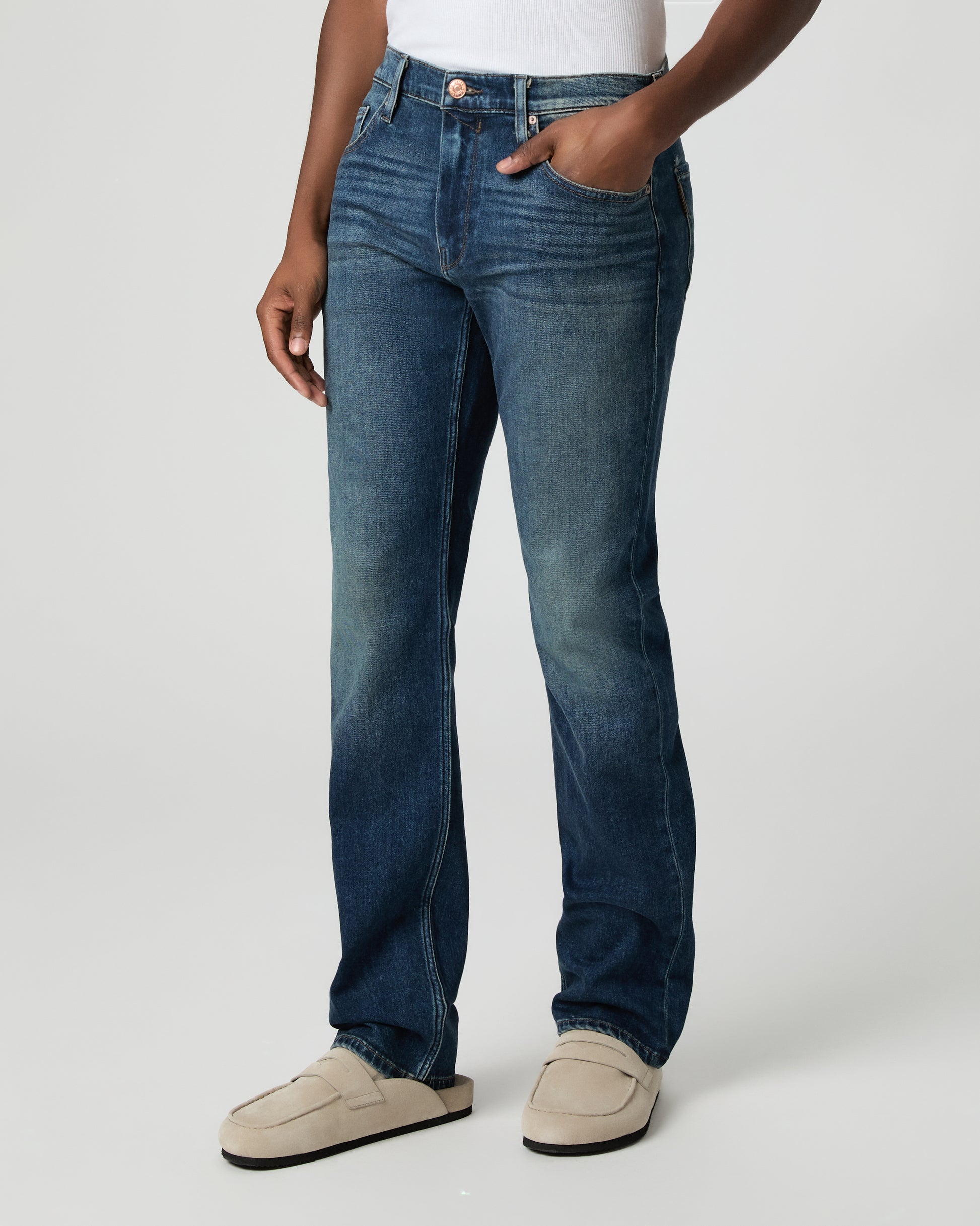 Man wearing a white shirt, blue jeans, and beige shoes on a plain background