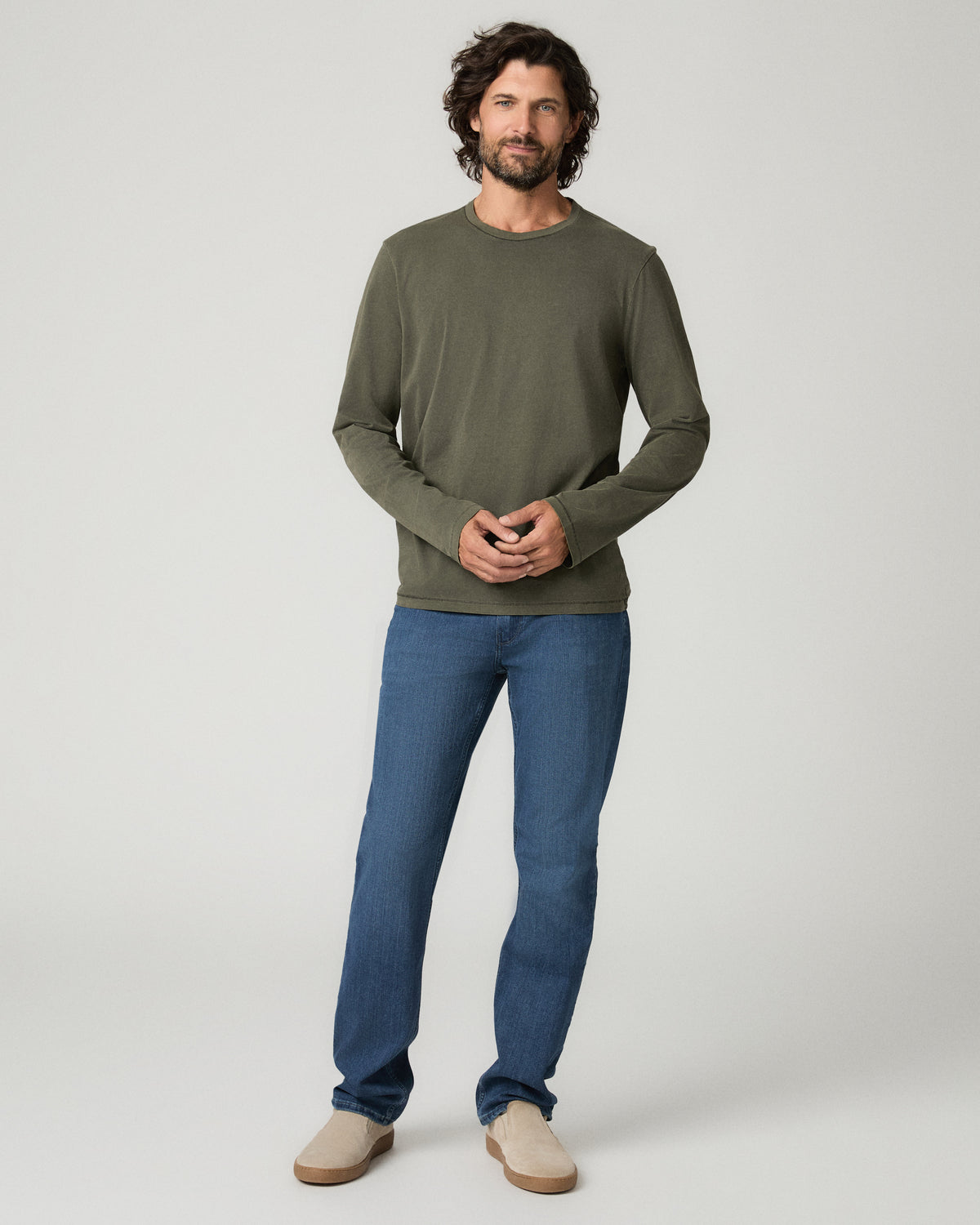 Full-length front view of a man wearing an olive green crew-neck sweater with hands clasped at his waist, paired with medium blue wash Elgin straight jeans and tan suede chukka boots.
