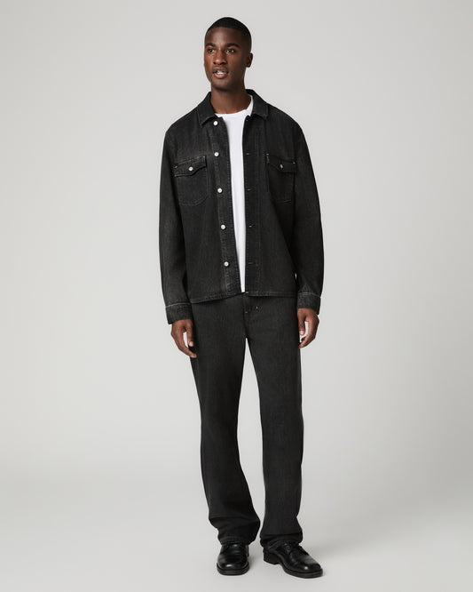 Full-length front view of a man wearing an open black denim trucker jacket over a white t-shirt, paired with washed charcoal-black Argo relaxed straight jeans and black shoes.