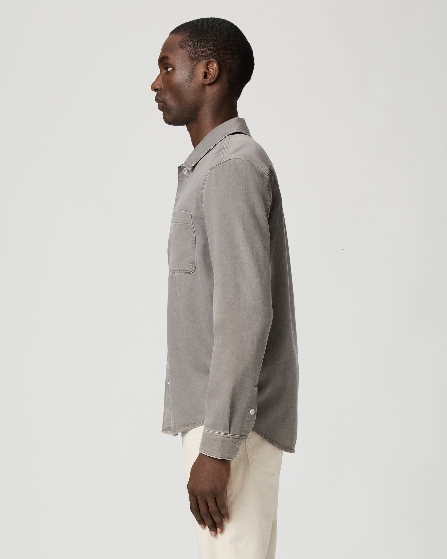 Man wearing a gray button-up shirt with a pocket and white pants on a white background