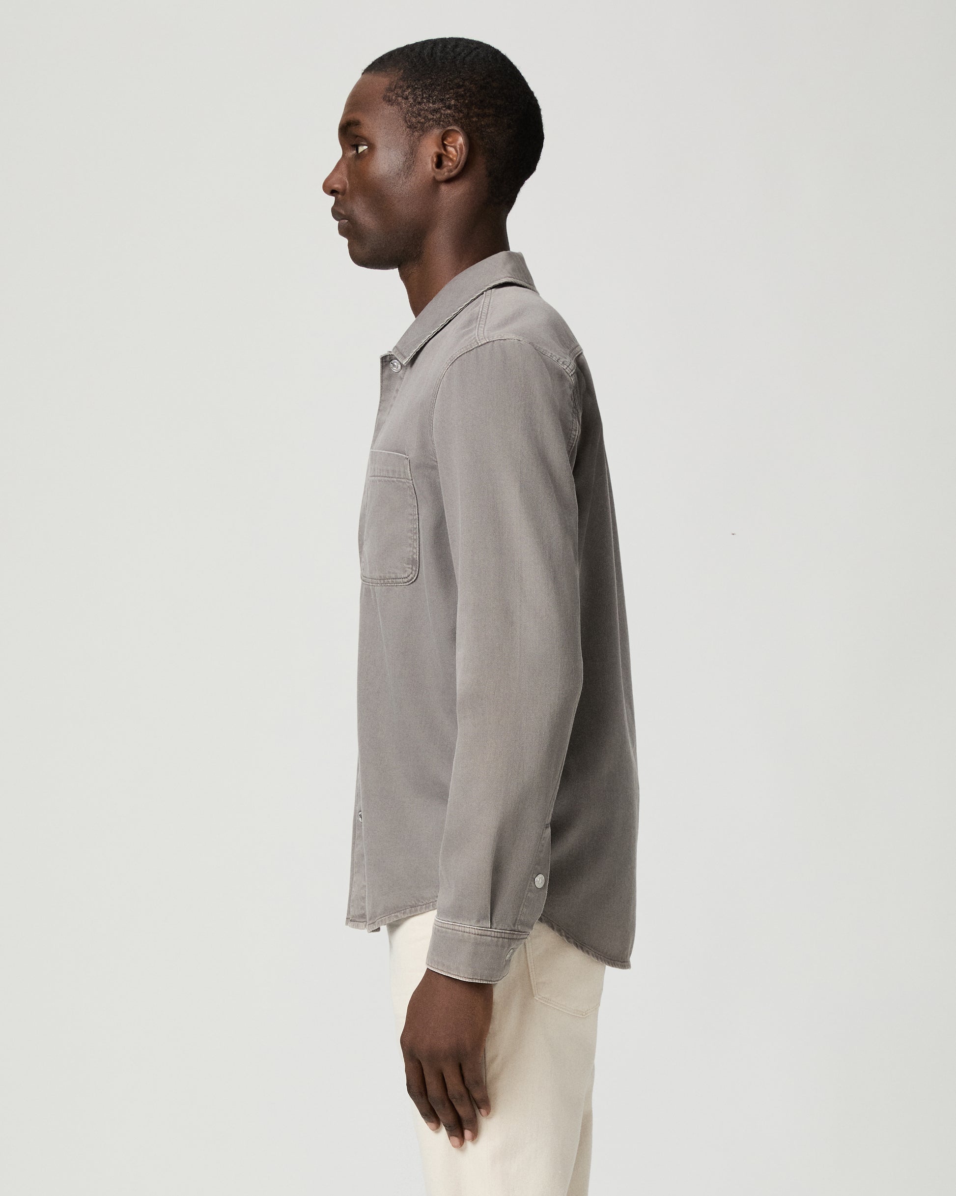 Man wearing a gray button-up shirt with a pocket and white pants on a white background