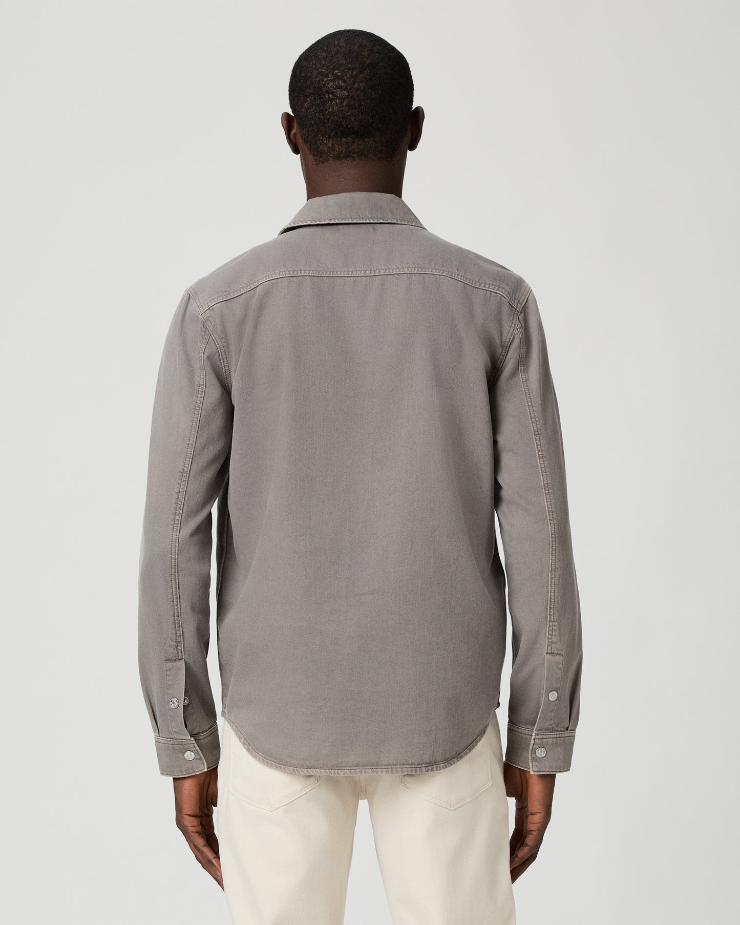 Man wearing a gray button-up shirt with a pocket and white pants on a white background