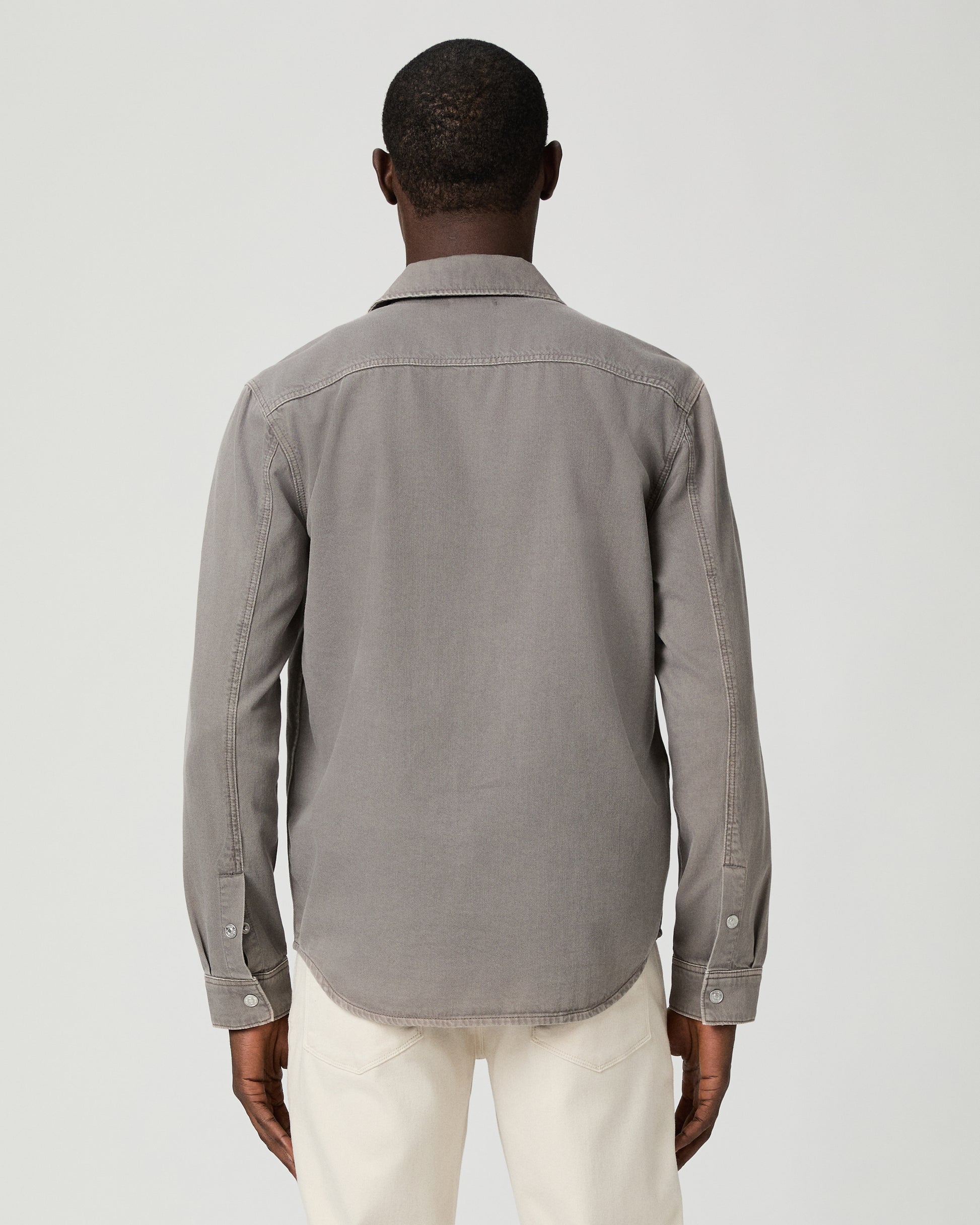 Man wearing a gray button-up shirt with a pocket and white pants on a white background
