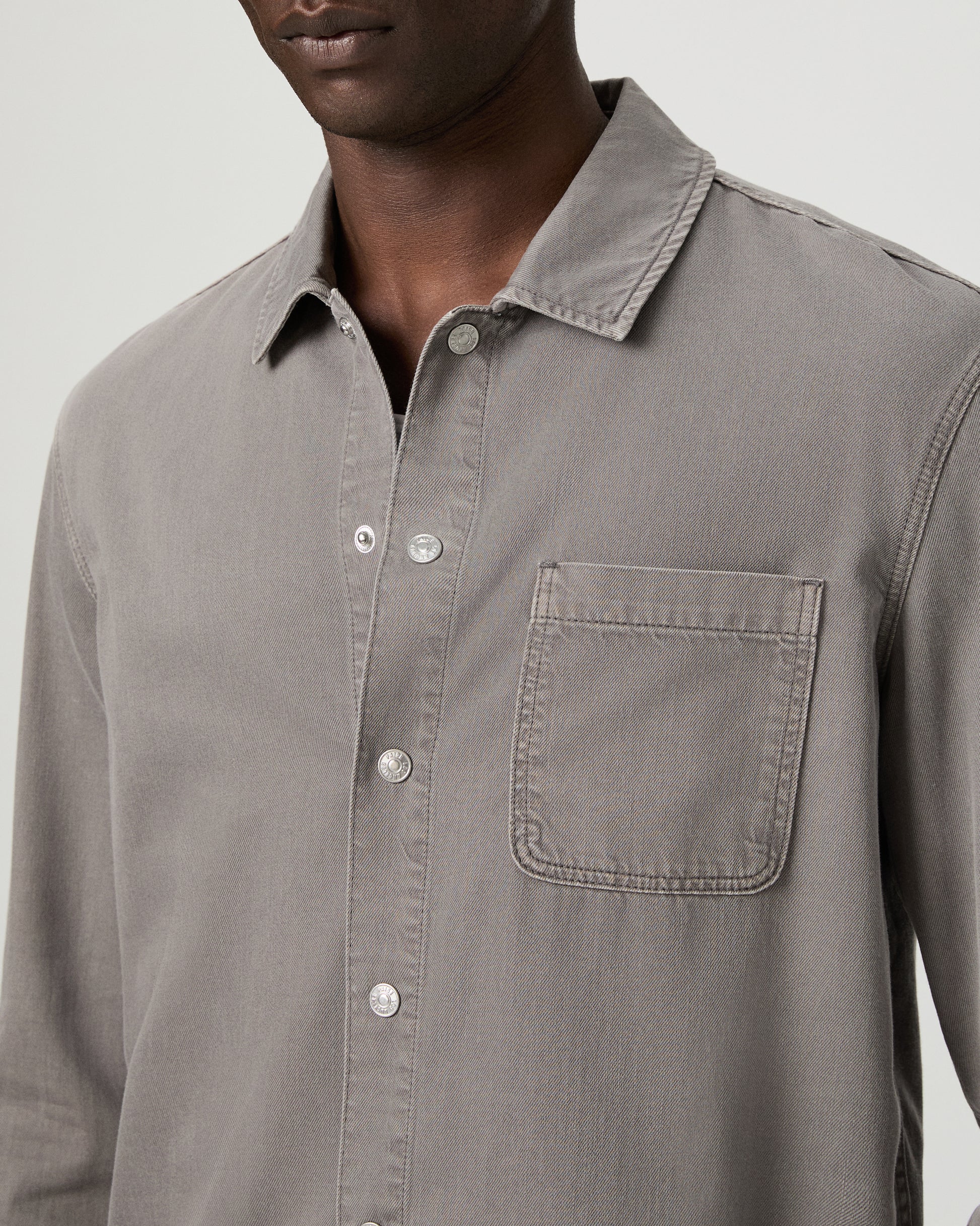 Man wearing a gray button-up shirt with a pocket and white pants on a white background