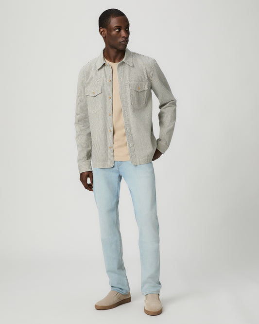 Man wearing a light gray flannel shirt, beige tee, light blue jeans, and brown shoes on a white background
