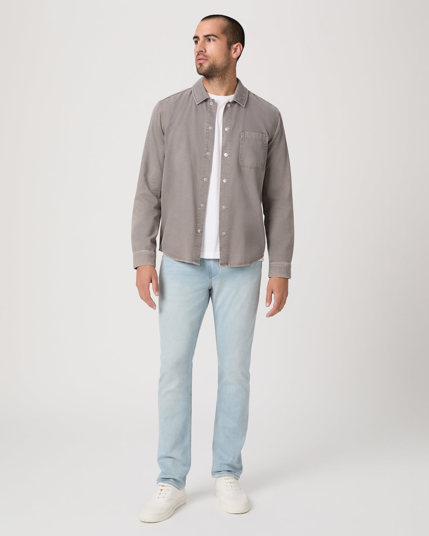 Man wearing a gray shirt over a white t-shirt, light blue jeans, and white sneakers on a white background