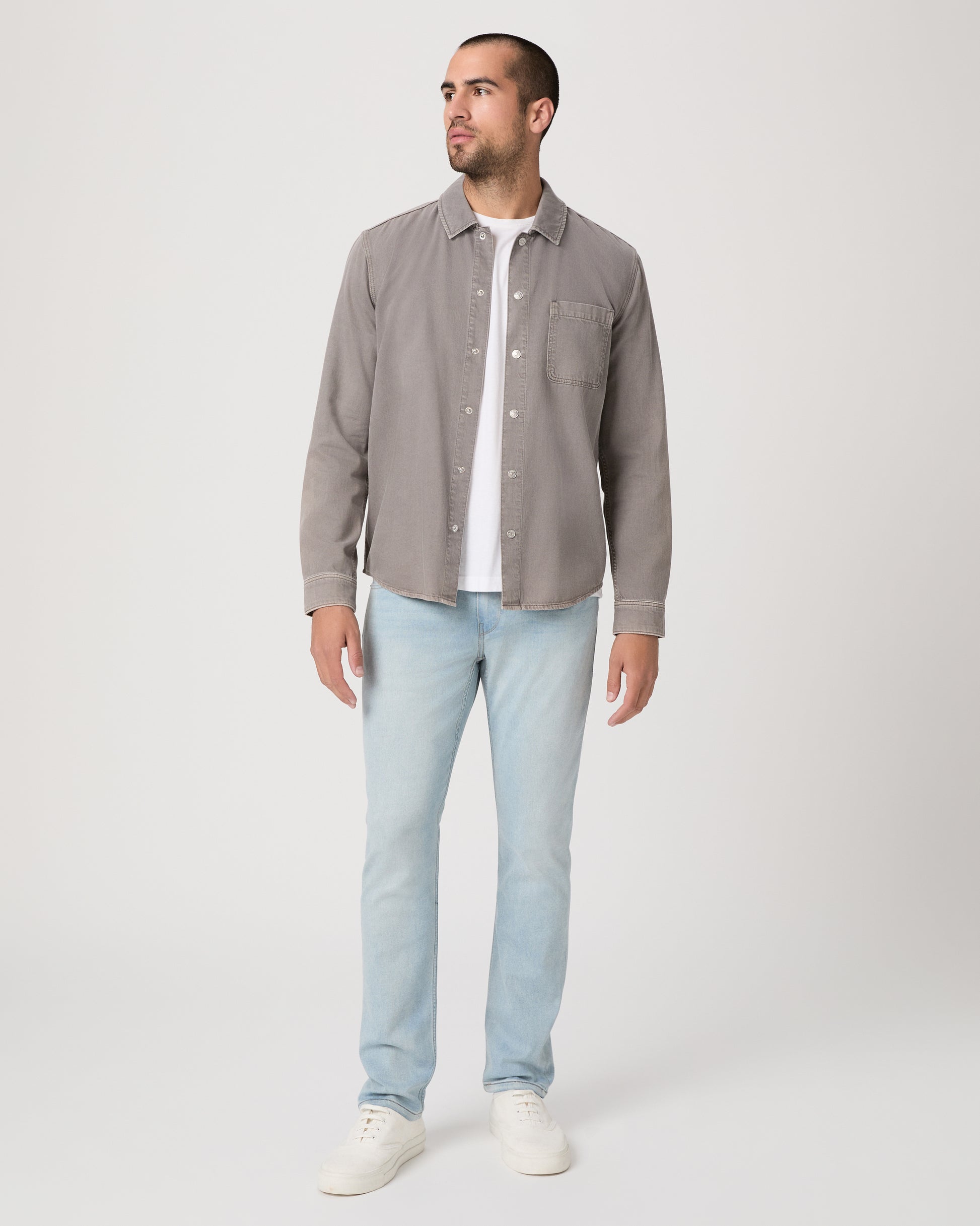 Man wearing a gray shirt over a white t-shirt, light blue jeans, and white sneakers on a white background