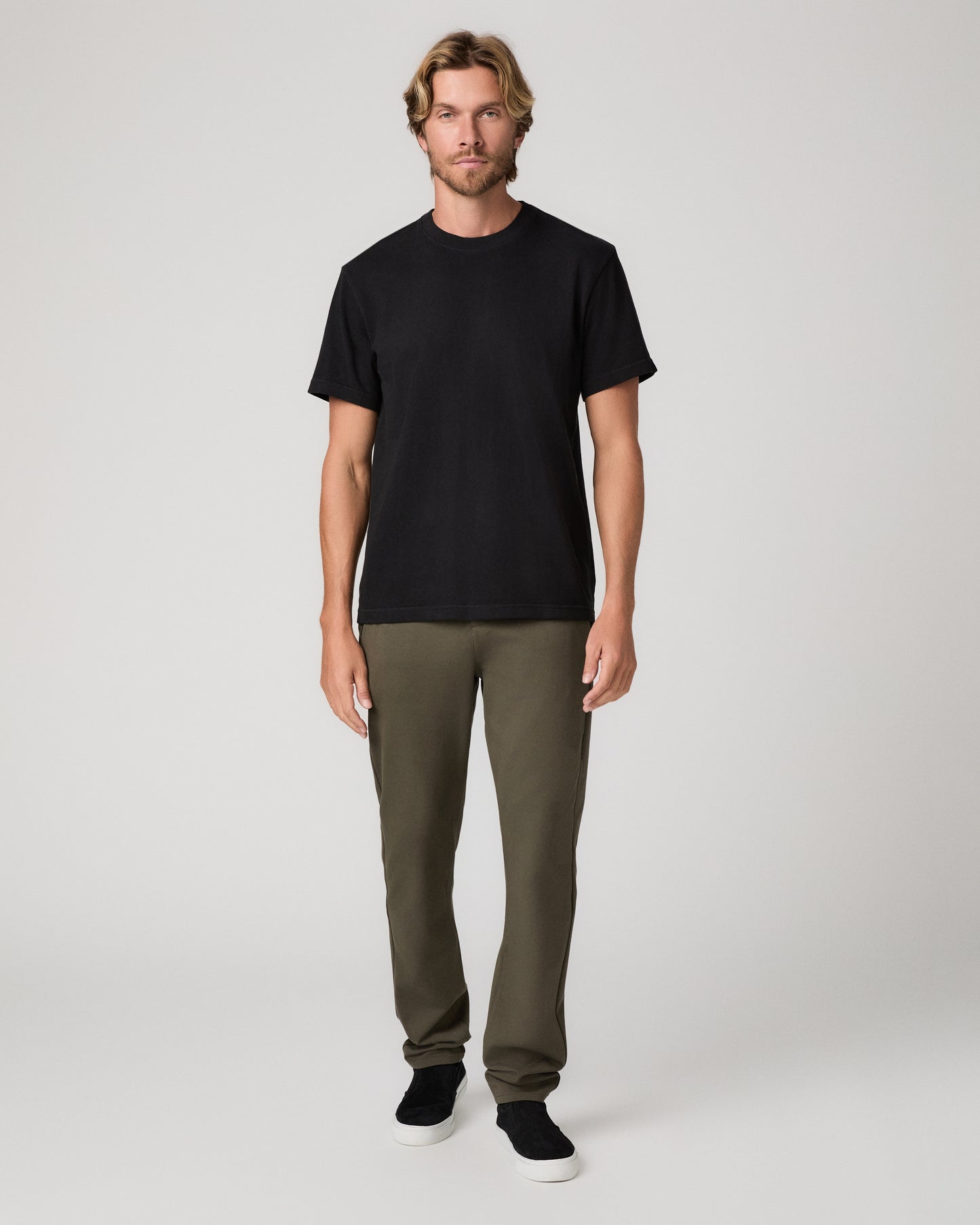 A man modeling a black short-sleeve crewneck t-shirt, olive green pants, and black slip-on shoes against a light gray studio background.