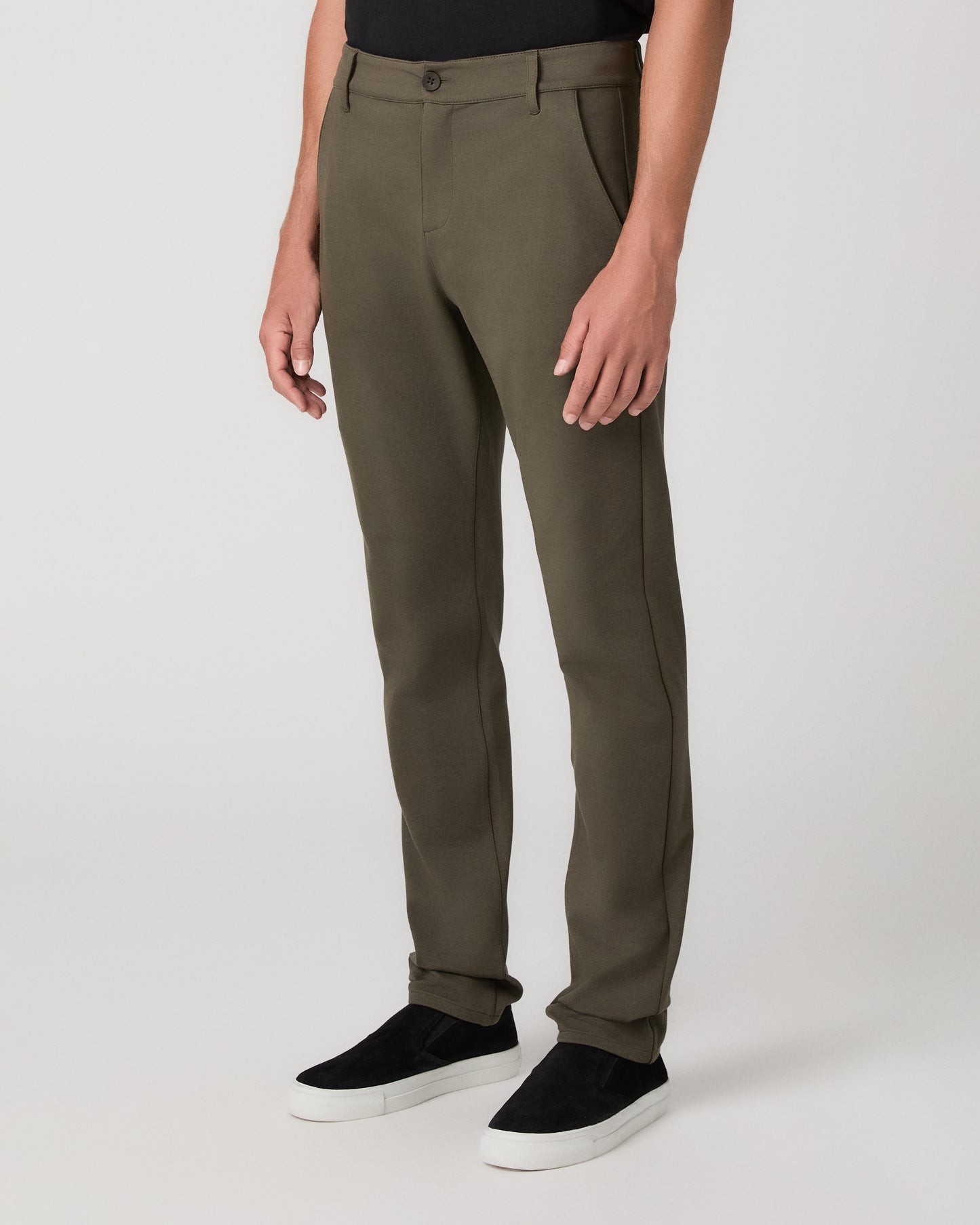 A man modeling a black short-sleeve crewneck t-shirt, olive green pants, and black slip-on shoes against a light gray studio background.