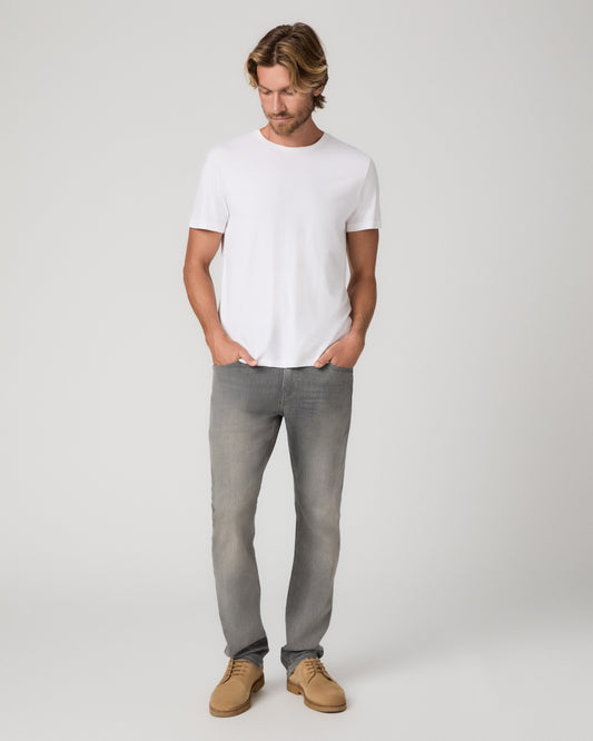 A man wearing a white crew-neck t-shirt, gray slim-fit jeans, and tan suede shoes, standing against a light gray studio background.