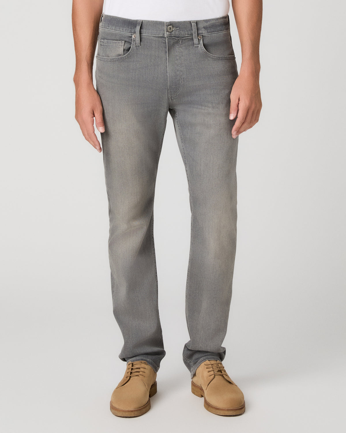 A man wearing a white crew-neck t-shirt, gray slim-fit jeans, and tan suede shoes, standing against a light gray studio background.
