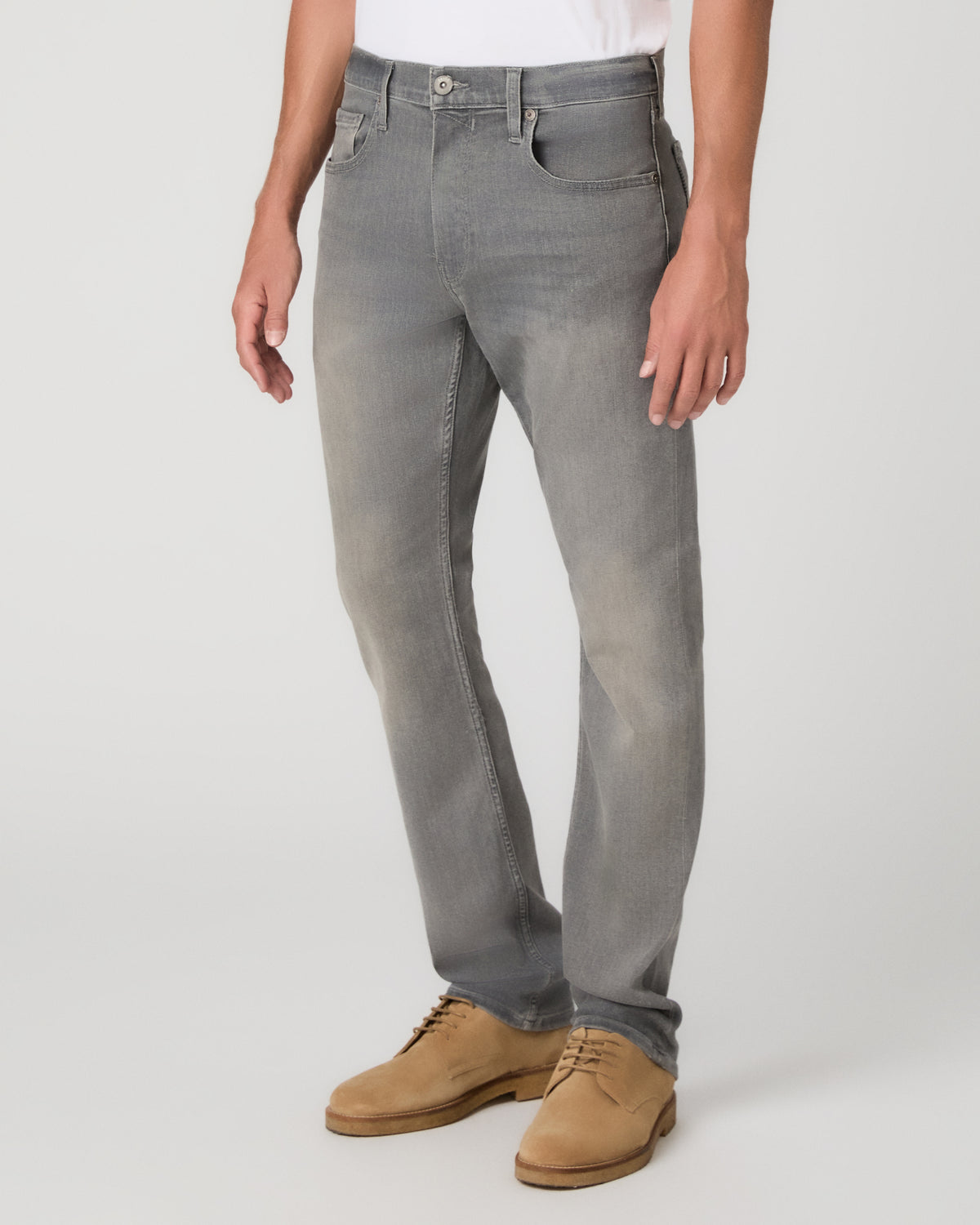 A man wearing a white crew-neck t-shirt, gray slim-fit jeans, and tan suede shoes, standing against a light gray studio background.