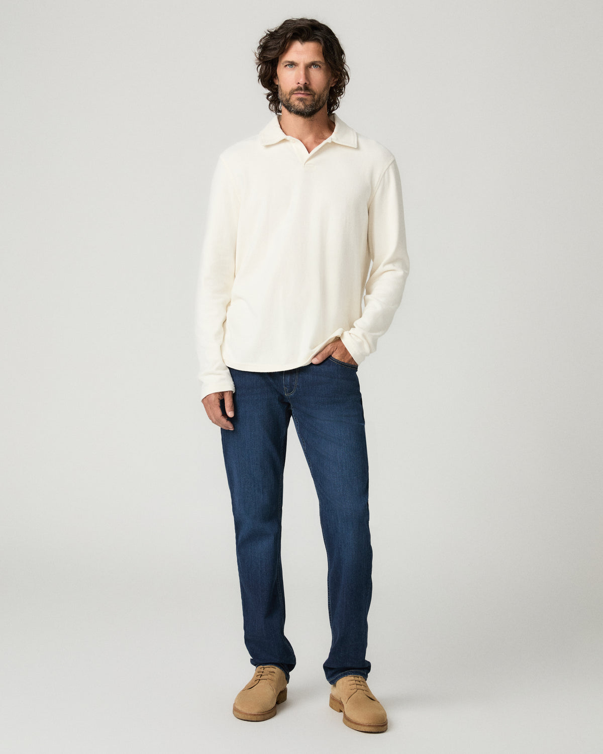 Full-length front view of a man with wavy dark hair wearing an off-white long-sleeve popover shirt with an open collar, one hand in his pocket, paired with deep blue slim-straight jeans and tan suede shoes.