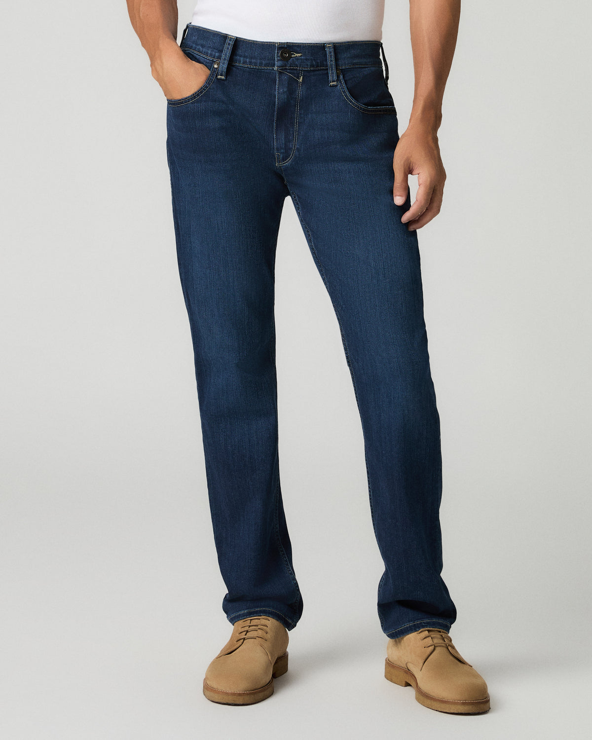 Close-up front view of a man with one hand in his pocket wearing deep blue slim-straight jeans, white t-shirt visible above the waistband.