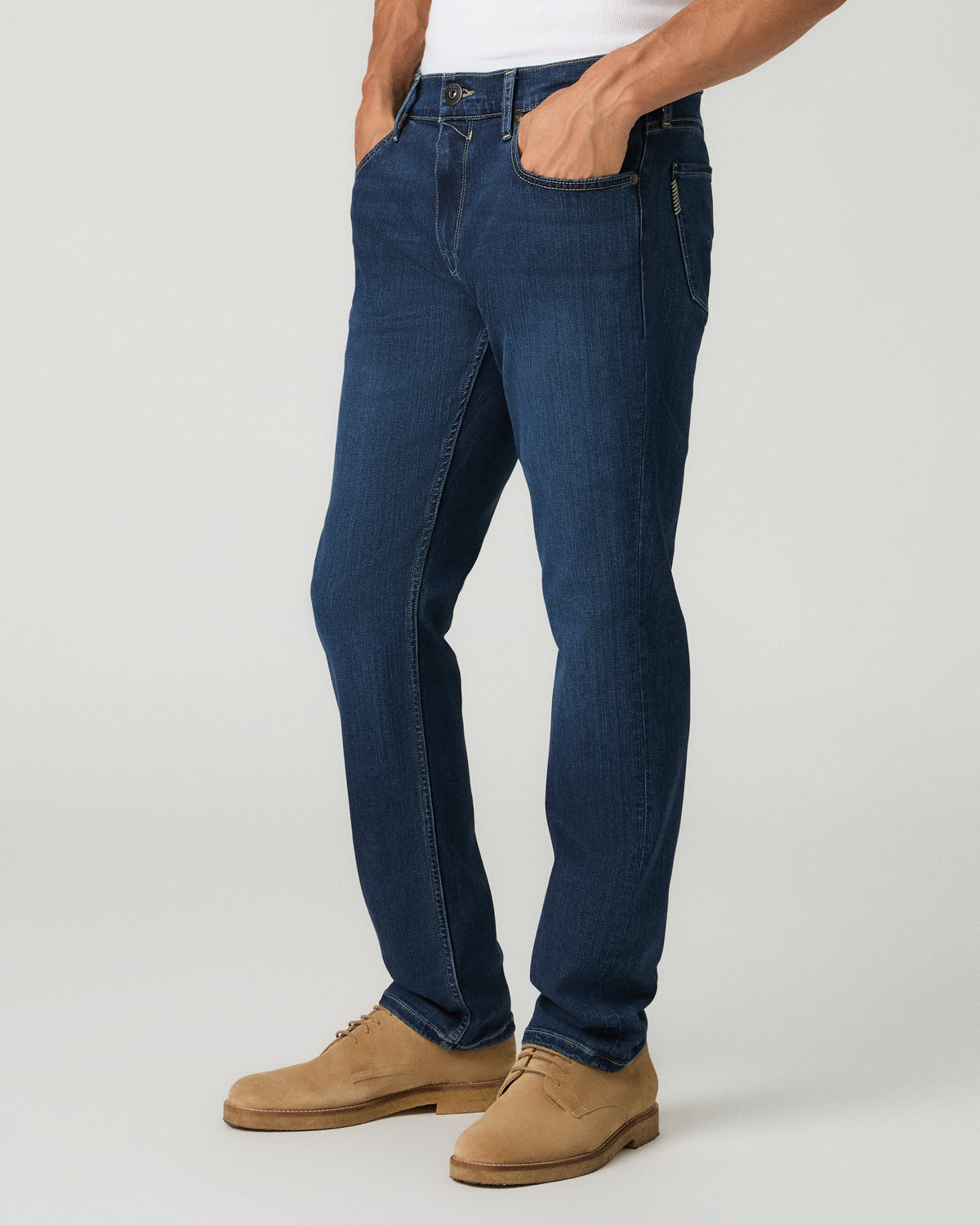 Three-quarter side view of a man with both hands in his pockets wearing deep blue slim-straight jeans and tan suede shoes.