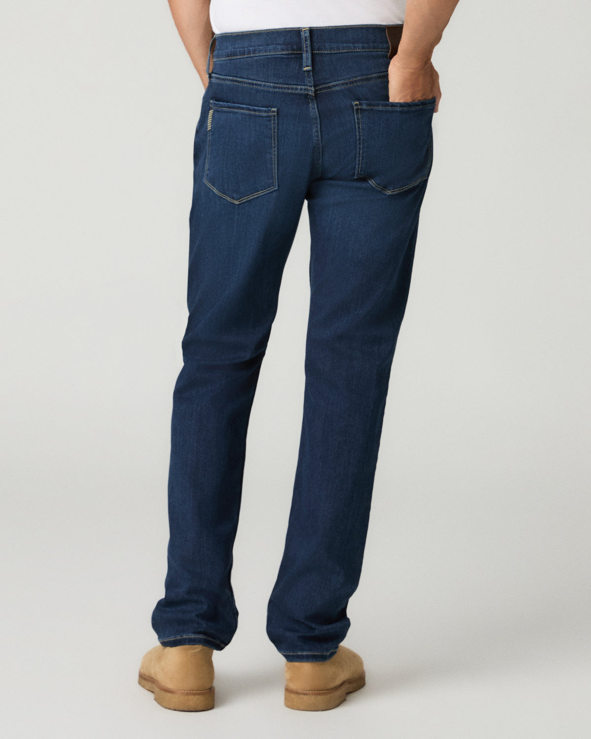 Full-length back view of a man with hands in his back pockets wearing deep blue slim-straight jeans and tan suede shoes.