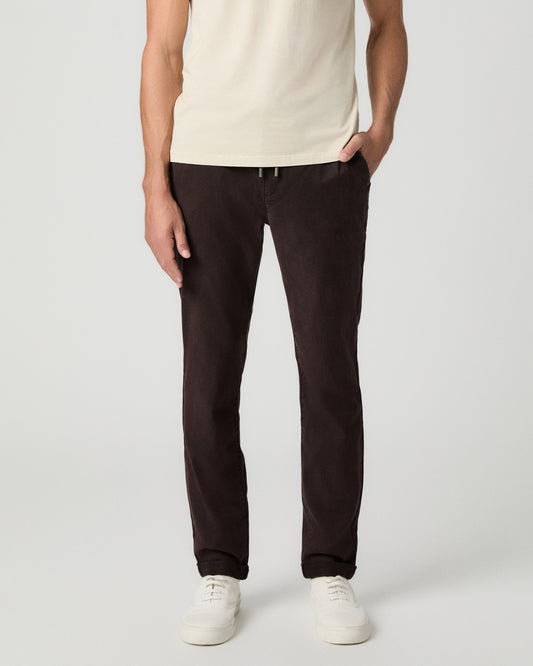 Front view from the waist down of the Fraser Pant in Mole, a dark brown drawstring pant, with one hand in the pocket, worn with a cream tee and white sneakers