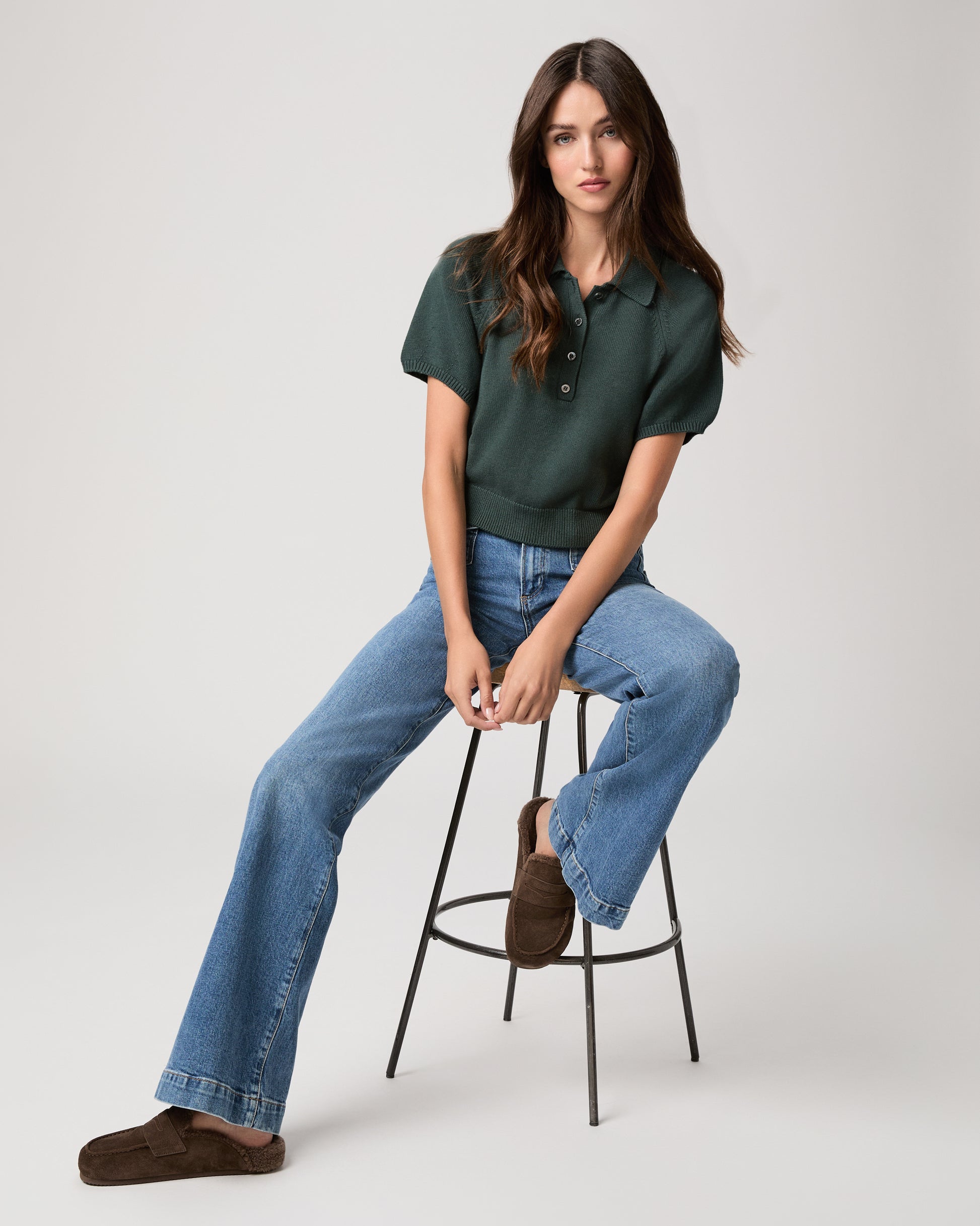 Woman sitting on a stool wearing a green top, medium wash jeans and chocolate color slipper with shearling lining on a light gray background