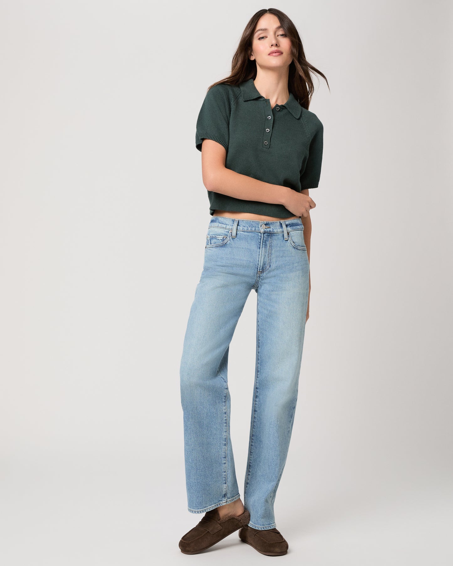 Woman wearing a green top, medium wash jeans and chocolate color slipper with shearling lining on a light gray background