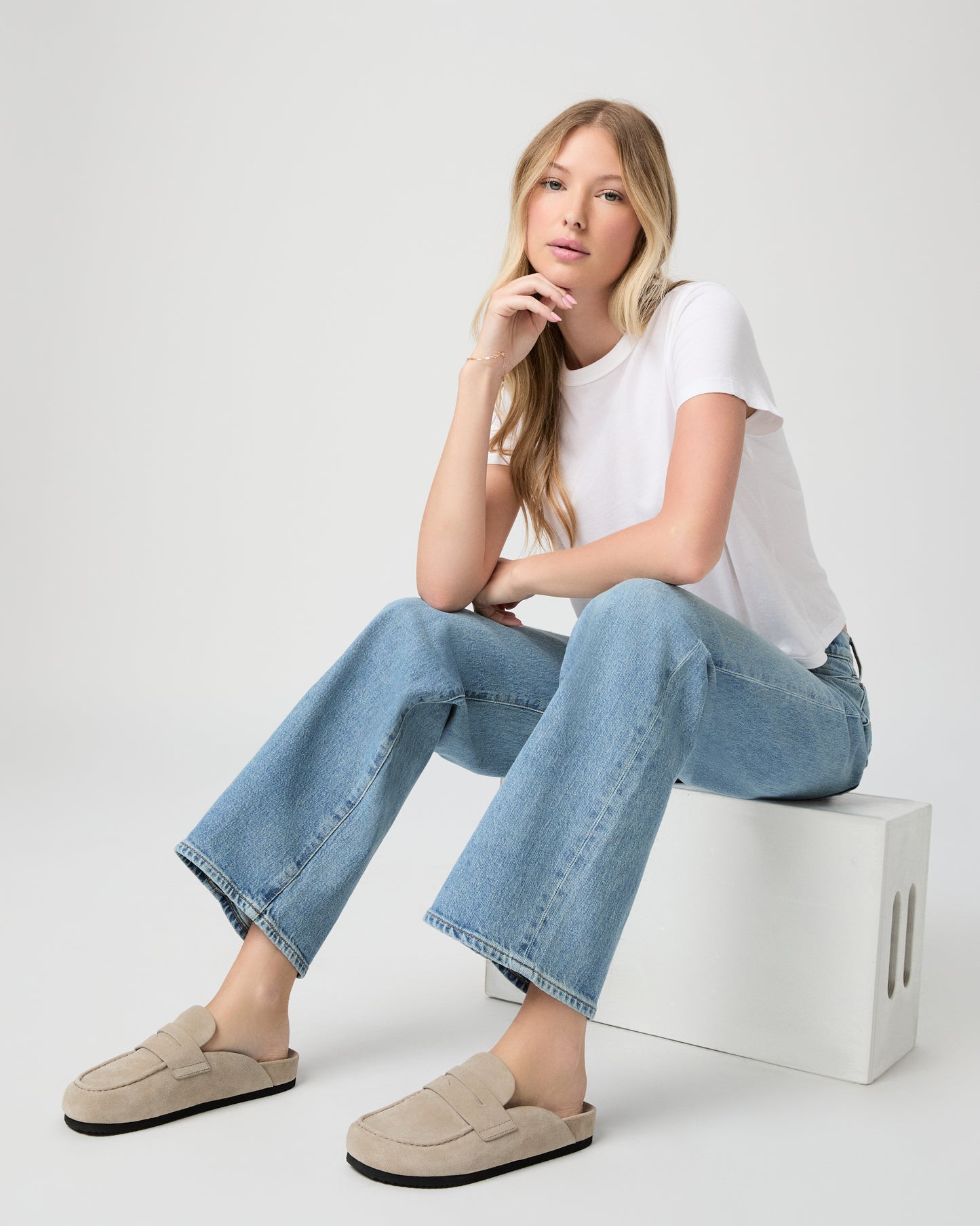 Women sitting down wearing white tee, blue jeans and beige slipper on a light gray background