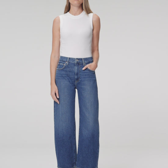 Woman wearing a white top and medium wash blue jeans paired with black boots on a plain background