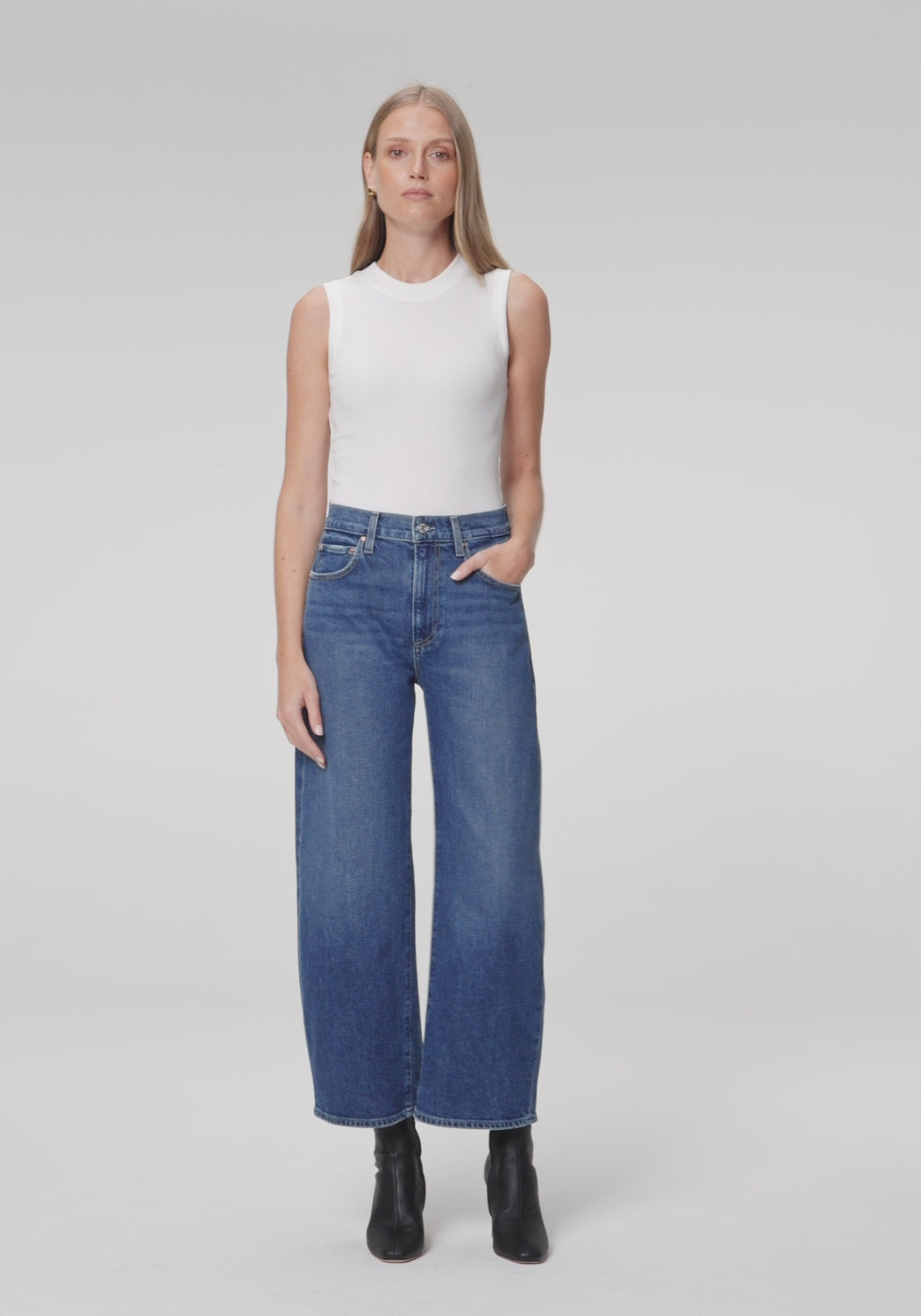 Woman wearing a white top and medium wash blue jeans paired with black boots on a plain background